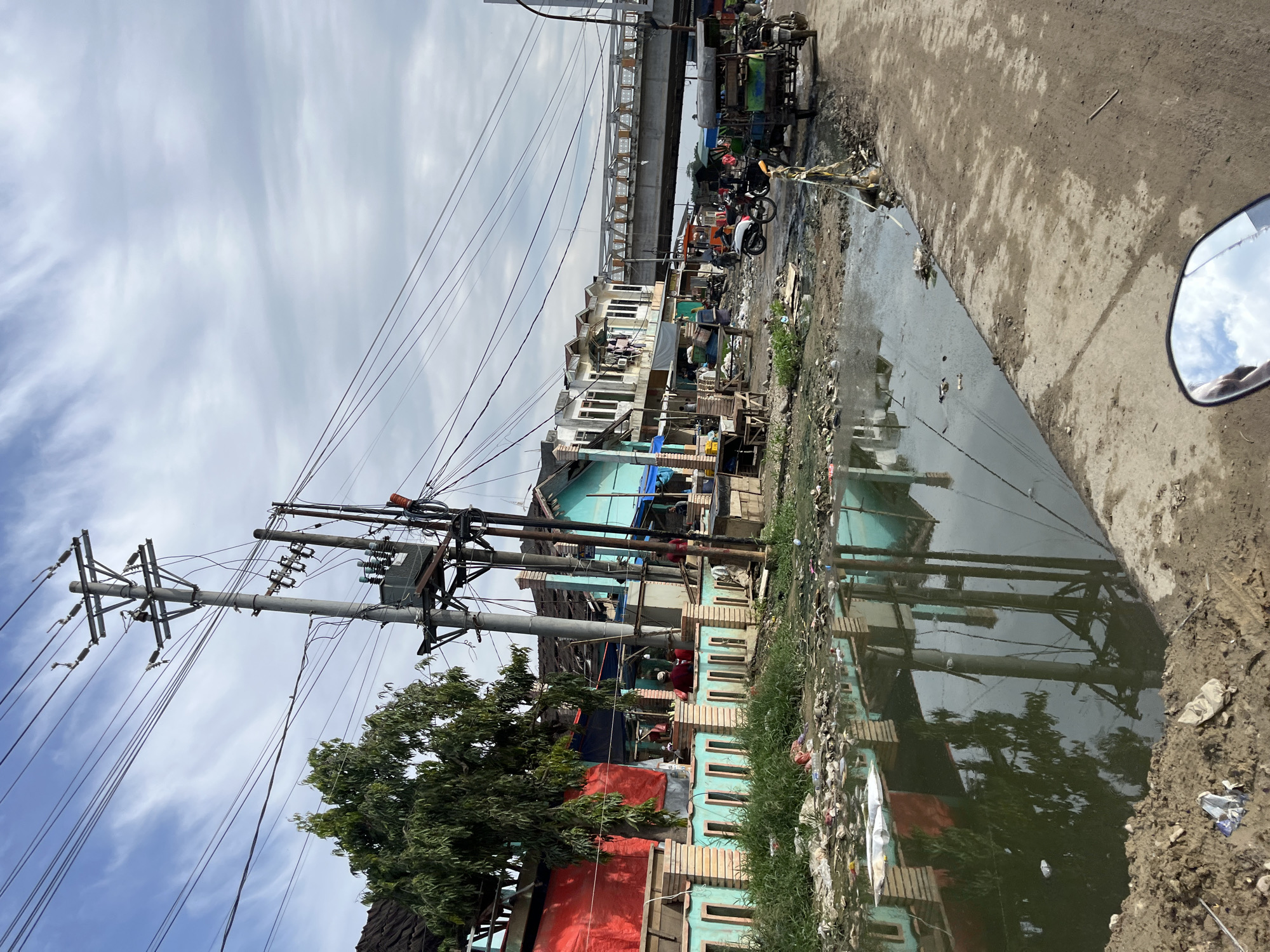 This image depicts a street scene in the city of Kota Kuala Simpang, Indonesia. A body of murky water, likely a drainage ditch or canal, runs along the left side of the road. This waterway is filled with debris and garbage, including plastic bags, bottles, and other refuse. Reflections of buildings and utility poles are visible on the water's surface.
Along the edge of the water, there are several buildings with colorful facades, including one painted red and another turquoise. A light-colored, decorative fence with vertical elements separates some of the buildings from the waterway. Lush green grass grows in patches along the embankment.
Utility poles with numerous electrical wires crisscross the sky. A transformer is mounted on one of the poles. Beyond the immediate buildings, a modern pedestrian bridge spans across the scene, indicating a more developed area further into the city.
The road itself appears to be unpaved or made of packed dirt and gravel, with some scattered debris. Several vehicles are present, including a three-wheeled vehicle (likely an auto-rickshaw or bemo) and a motorcycle. A few individuals can be seen near the buildings and vehicles. The sky is partly cloudy with a mix of blue sky and white clouds. The overall impression is of a somewhat neglected urban environment.