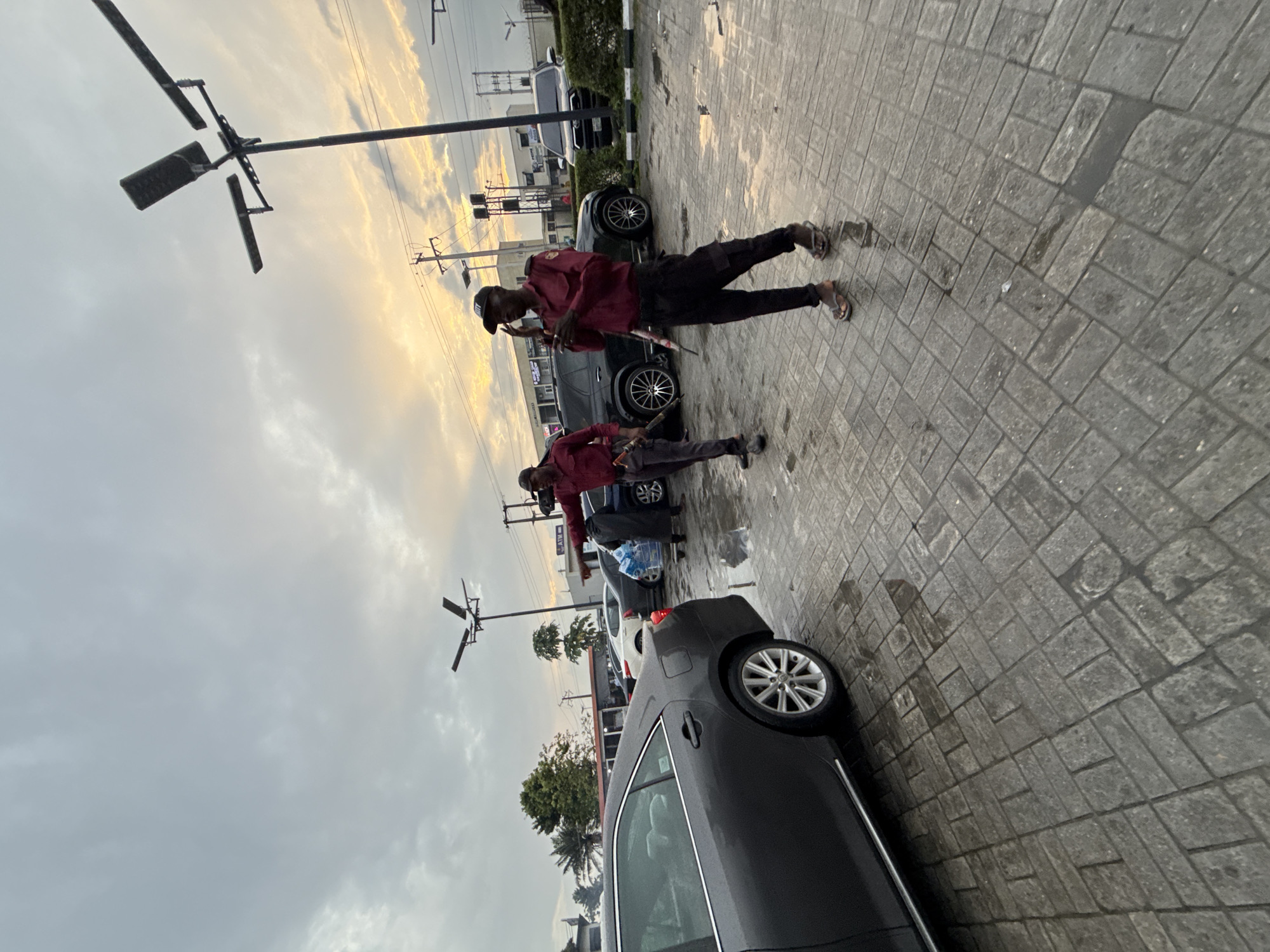 In Ado, Nigeria, two uniformed security guards stand on a wet, paved lot, possibly a car park, under a cloudy sky with hints of sunset. The guard on the left, wearing a maroon shirt and dark pants, holds a long, baton-like object. The guard on the right, similarly dressed, is speaking into a handheld device. Several cars are parked in the background, and a person is pushing a shopping cart. Streetlights and power lines are visible in the sky. The overall scene suggests a busy commercial area in Ado, Nigeria, potentially experiencing or having recently experienced rain, given the wet ground.