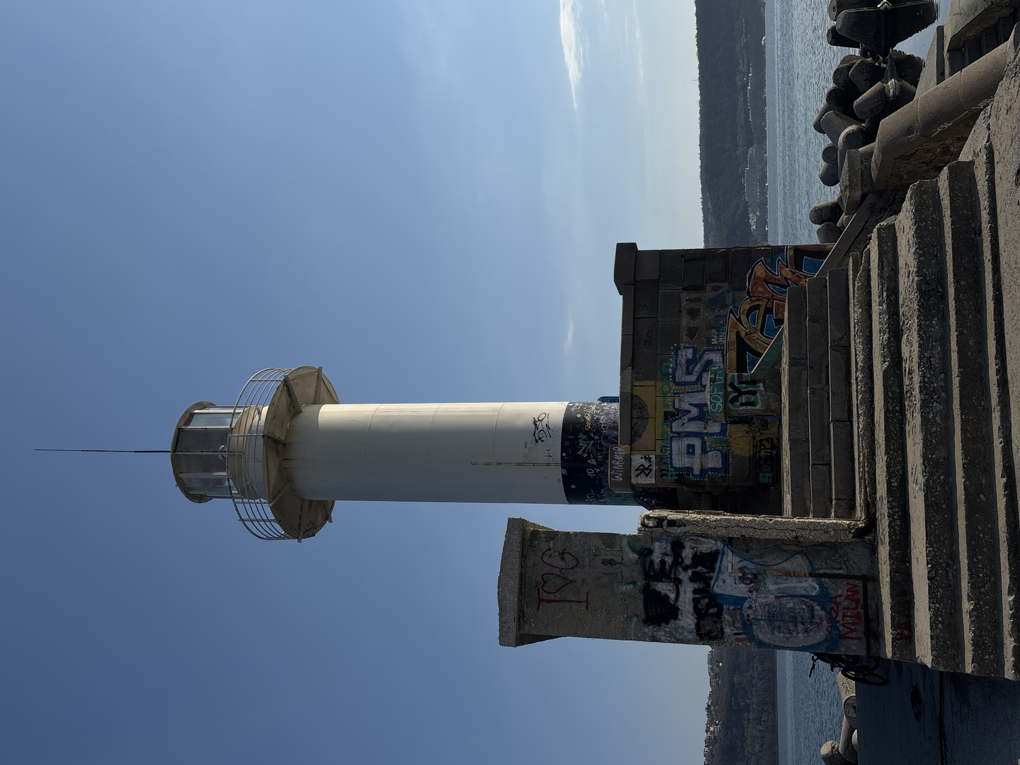 The image depicts a view of the Varna Lighthouse in Varna, Bulgaria, set against a clear blue sky. The lighthouse is a cylindrical white structure with a black band near its base and a metal cage surrounding its lantern room. Graffiti covers the concrete structures around the base of the lighthouse. A set of concrete stairs leads up alongside the lighthouse base. In the background, the sea and a stretch of land are visible. The lighthouse appears to be situated on a concrete pier or breakwater. The photo is taken from a low angle, looking upwards.