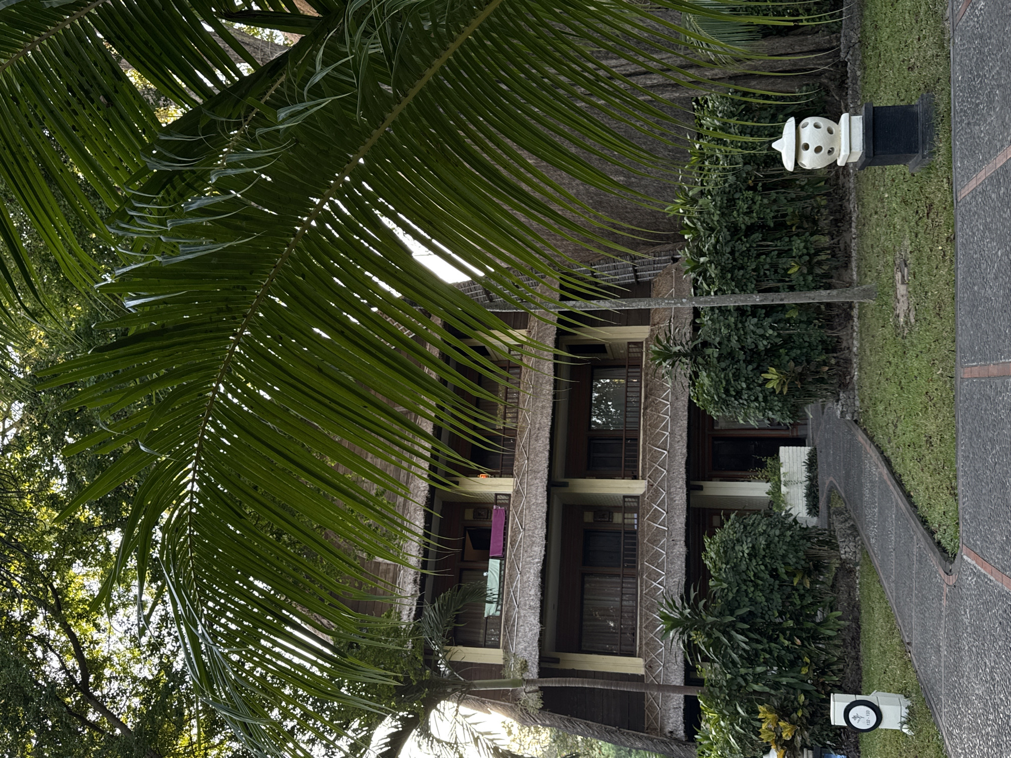 The image captures a tranquil scene in Denpasar, Indonesia, likely at a resort or hotel. In the foreground, a pathway made of grey cobblestones curves through a lush green lawn, bordered by manicured bushes and palm trees. The path leads towards a multi-story building with a distinctive thatched roof and balconies. The building's facade is a warm beige, with dark wooden accents around the windows and balconies. Two colorful towels, one green and one purple, are draped over a balcony railing on the second floor. On the right side of the image, a white, ornamental lantern sits atop a dark rectangular base on the grass. A tall, slender tree with a textured bark stands behind the lantern, partially obscured by the fronds of a large palm tree that dominates the upper right portion of the frame. The setting suggests a tropical atmosphere, with the natural elements harmonizing with the architecture. A white cylindrical marker with black lettering and a circular emblem is visible on the left side of the path.