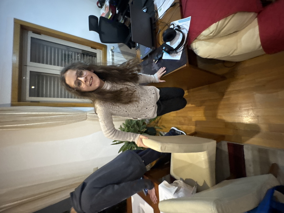 An indoor scene in Mirandela, Portugal, depicts two individuals within a furnished room. The primary subject, a young female with long brown hair and glasses, is positioned centrally, smiling directly at the camera. She wears a light-colored knitted turtleneck sweater and dark pants, and appears to be suspended mid-air with her feet off the polished wooden floor. Her left hand rests on the back of a light-colored upholstered chair, and her right hand is placed on a brown wooden desk to her right. To the left, a male figure is partially visible from the back, leaning over a separate wooden table covered with white papers. He wears a dark blue hooded sweatshirt and dark pants.
The room features a large window with a wooden frame and light-colored curtains in the background. Below the window, a white radiator is visible. To the right of the window is a black office chair. The wooden desk, where the female subject's right hand rests, holds an open black laptop, a black computer mouse, a black headset, and several white papers. In the foreground to the right, a section of a light-colored sofa or armchair draped with a red blanket is visible. A green potted plant is situated between the two tables. The floor is composed of polished wooden planks, and a striped rug is partially visible under the chair to the left. Overhead lighting casts shadows onto the floor.