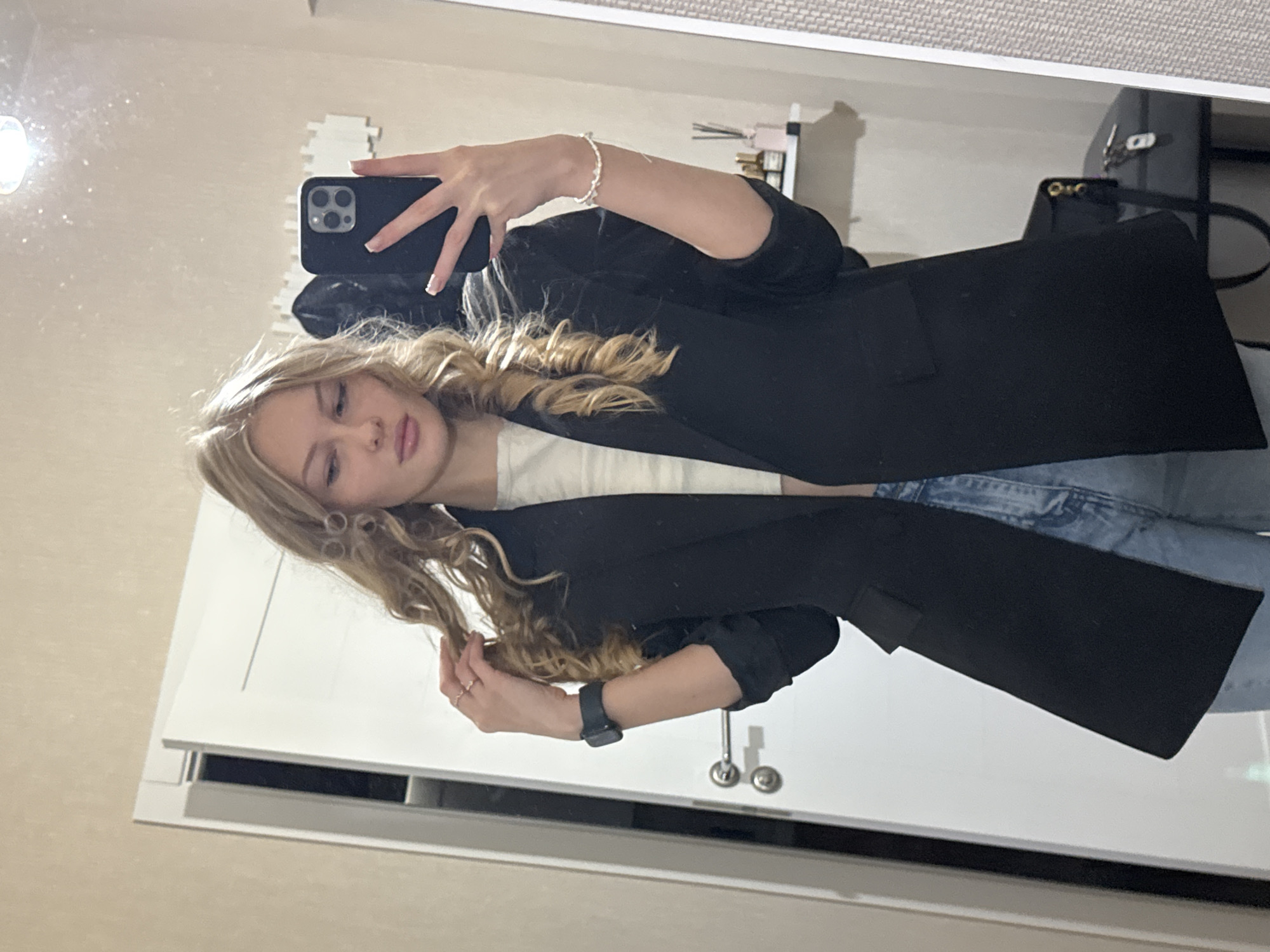 In an indoor setting, possibly a residential apartment in Perm, Russia, a young woman with blonde, wavy hair is taking a mirror selfie. She is wearing a black blazer over a cream-colored top and light blue jeans. Her left hand is touching her hair, and her right hand is holding a smartphone. She is wearing rings on her left hand, a watch on her left wrist, and a bracelet on her right wrist. The background includes a white door with a silver handle, a mirror, and a shelf with small bottles. The lighting suggests artificial light sources within the room.