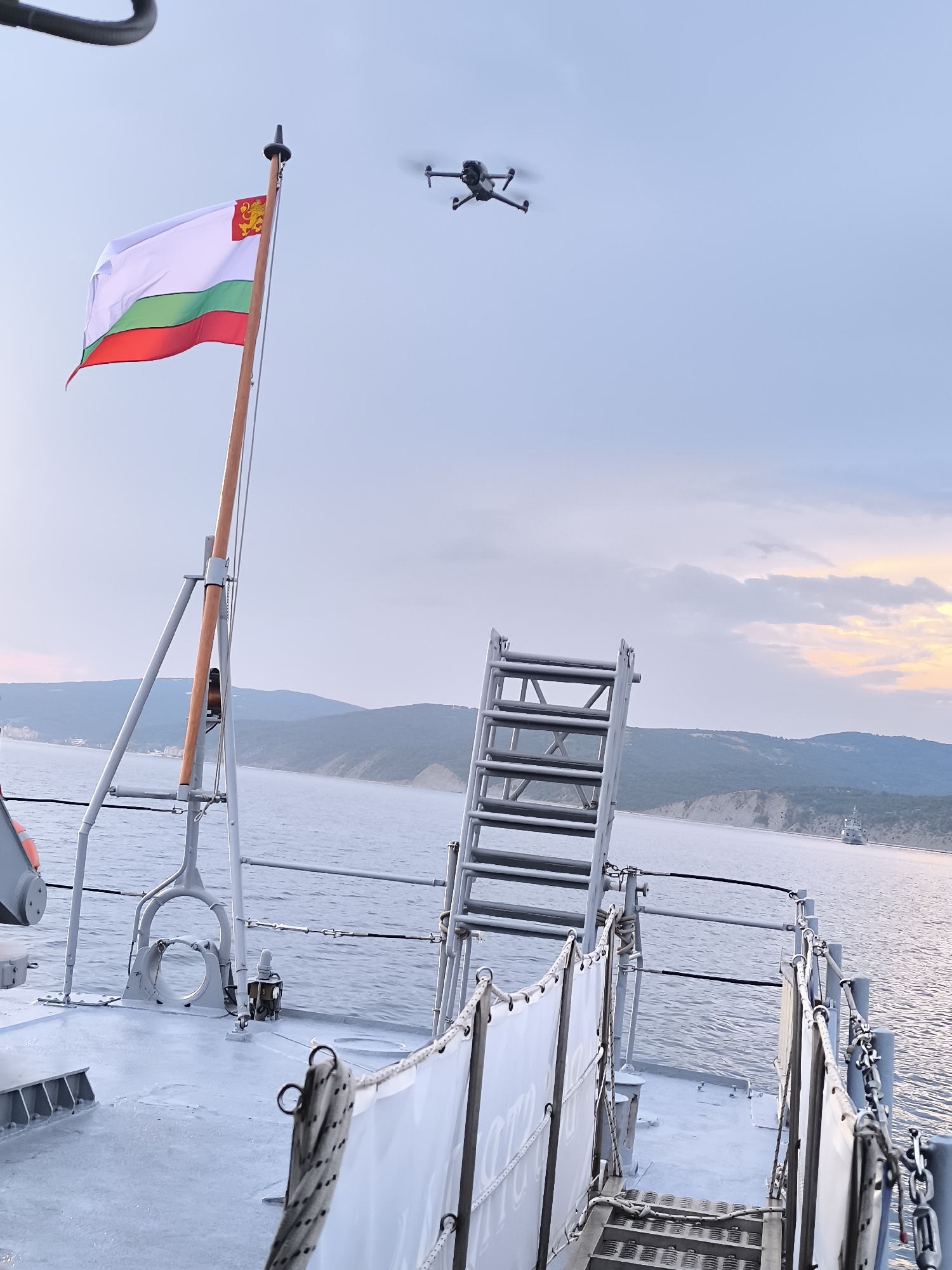 The image captures a view from the deck of a vessel at sea, likely near the coast of Emona, Bulgaria, given the presence of the Bulgarian flag. A drone is visible flying in the clear sky. In the foreground, part of the ship's deck is visible, including a ladder, railings, and a white banner. The Bulgarian flag, featuring white, green, and red stripes with a coat of arms on the white stripe, is prominently displayed on a flagpole. The background shows a coastline with hills and a distant ship on the water. The lighting suggests it is either dawn or dusk, with soft, diffused light illuminating the scene and a hint of color in the sky.