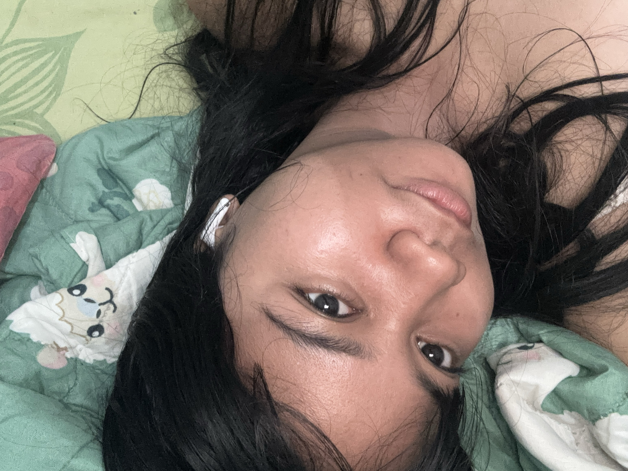 The image shows a close-up, overhead view of a person lying down on a bed with green and white patterned bedding. The person has long, dark hair spread around their face. They are wearing white earbuds. The lighting is soft, suggesting an indoor setting, possibly a bedroom. The background consists of the patterned sheets and pillow. There is no indication of the specific location within North Jakarta, Indonesia.
