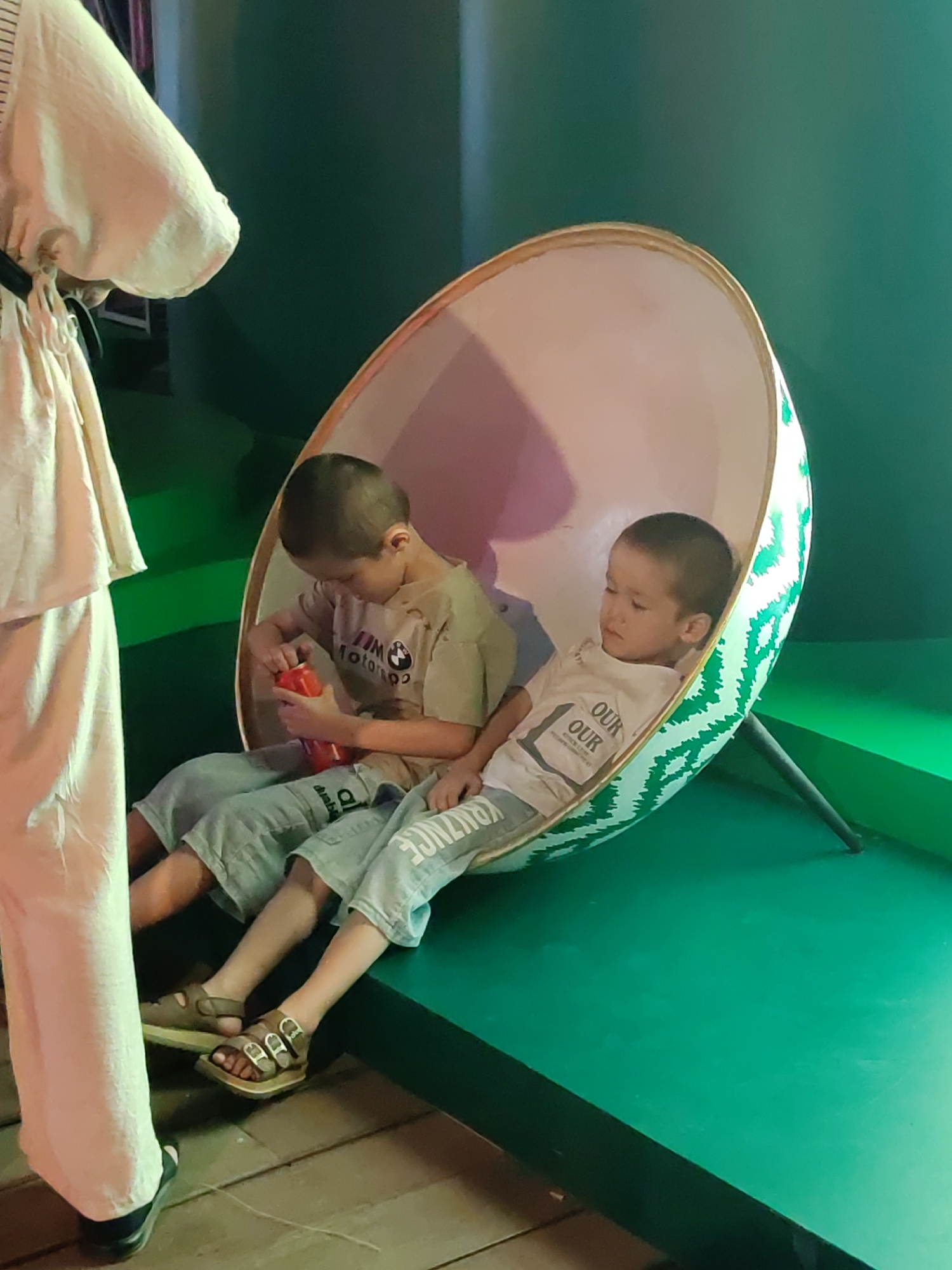 Two young boys are sitting inside a large, egg-shaped chair with a green and white striped pattern on the outside. The chair is tilted, with its base resting on a dark-colored stand. The boy on the left is wearing a light brown t-shirt with a BMW logo and grey sweatpants. He is holding a red object in his hands. The boy on the right is wearing a white t-shirt with "OUR OUR" printed on it in black letters, and also grey sweatpants. Both boys are looking downwards. To the left of the chair, a person is standing, partially visible, wearing a long, loose-fitting light-colored garment. The background is a solid dark green wall. The floor beneath the chair is made of wooden planks, while the area in front of the chair appears to be a green platform. The scene suggests an indoor setting, possibly an exhibition or a public space in Tashkent, Uzbekistan, given the presence of the distinctive egg-shaped chair often found in themed attractions or play areas.