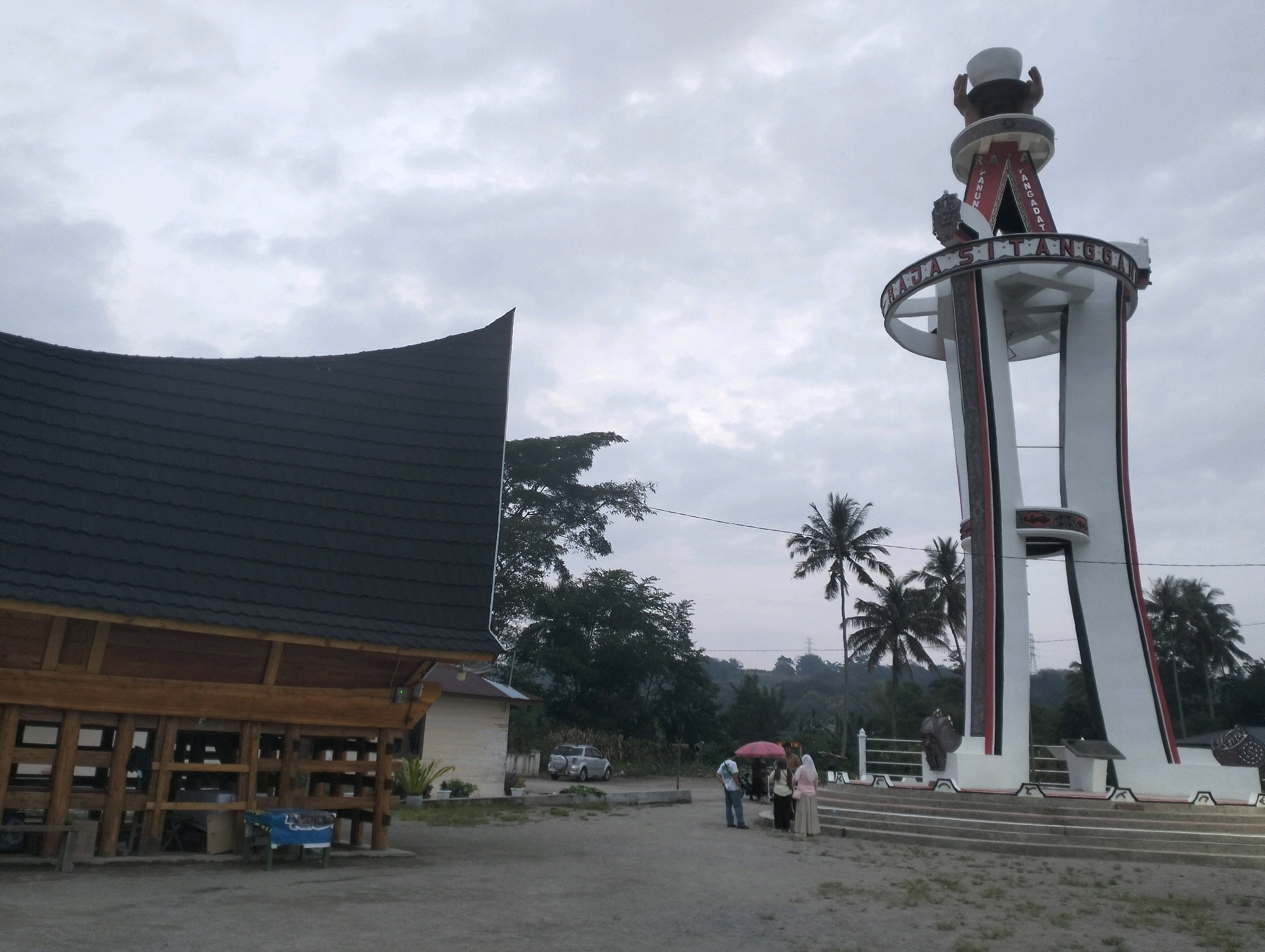 The image depicts an outdoor scene with a large, white monument on the right, featuring red and black accents and what appears to be a depiction of hands holding a bowl at the top. The monument has text inscribed on it, partially visible as "PANGADAT" and "RAJA SITANGGA". To the left of the monument is a building with a distinctive dark, curved roof, constructed with visible wooden beams. In the foreground, several people are gathered, some holding an umbrella. A silver SUV is parked behind the building. Palm trees and other foliage are visible in the background, suggesting a tropical or subtropical environment. The sky is overcast with clouds. The scene appears to be a public space, possibly a historical site or landmark in the city of Parlondut, Indonesia.