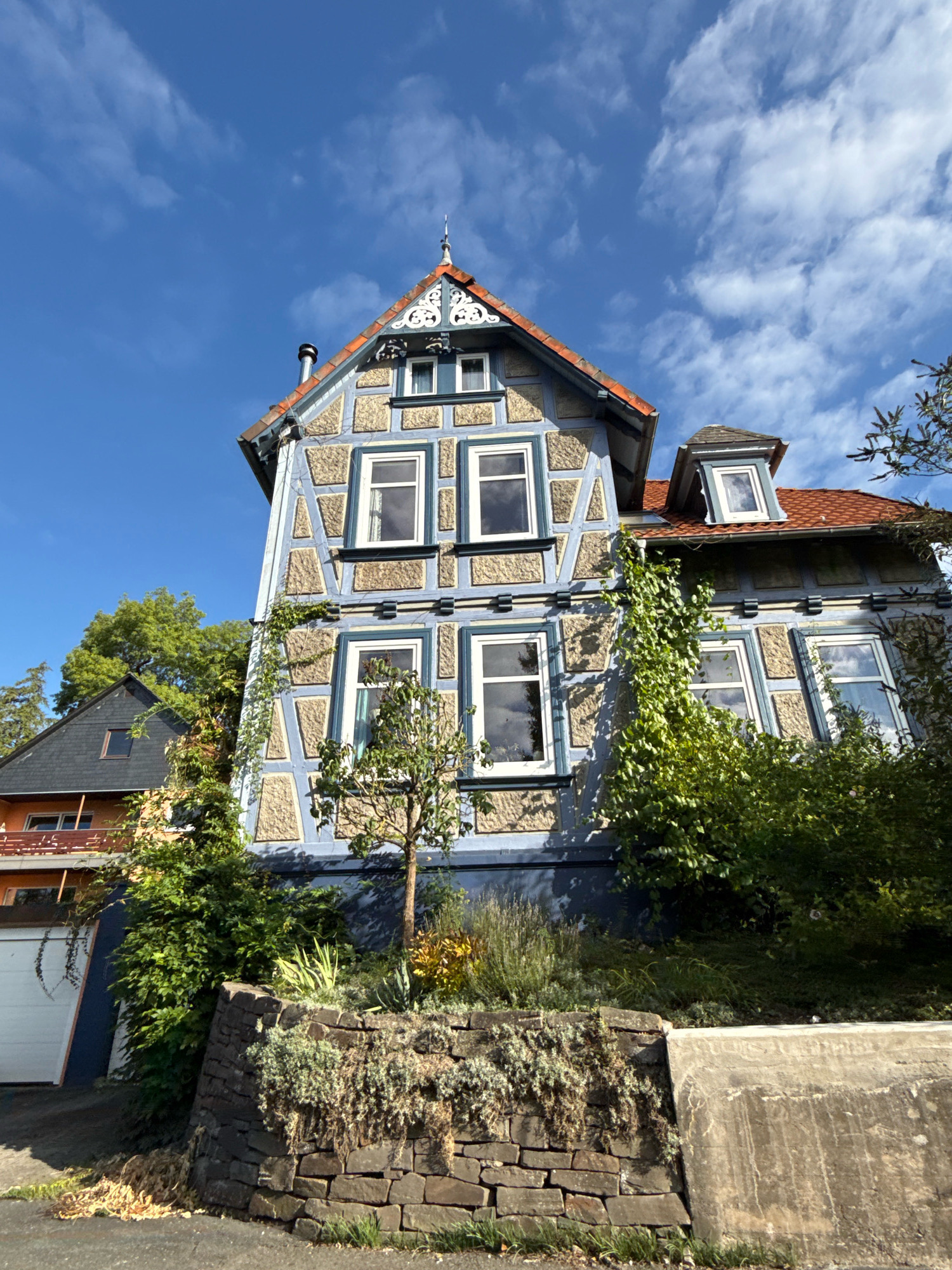 The image captures a low-angle view of a traditional half-timbered house in Osterode am Harz, Germany. The house features a light blue facade with beige timber framing and a red tiled roof. Multiple windows with white frames are visible on the front of the house. Green ivy is growing up the side of the building. In front of the house, there is a stone wall retaining a sloped garden area with various plants and shrubs. A driveway with a white garage door is partially visible on the left side of the image. The sky above is blue with scattered white clouds. The scene appears to be a residential area on a clear day.