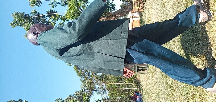 An outdoor, brightly lit, full-body shot captures an older man, likely of African descent, standing in Burundu, Kenya. He is positioned towards the left of the frame, with his body slightly angled and his bald head turned to face the right. The man is wearing a dark green jacket or coat over a dark blue shirt and dark blue trousers, paired with dark sneakers with light soles. His right hand is visible, open and relaxed.
The setting is a grassy field under a clear, bright blue sky, indicating sunny weather. Several trees, featuring green foliage, line the background, with some taller, slender trees extending upwards. The ground cover consists of green grass with some drier, brown patches. Strong shadows are cast, suggesting it is either mid-morning or early afternoon. In the distant background, faint shapes hint at other people or structures within the natural landscape. There is no visible text in the image.