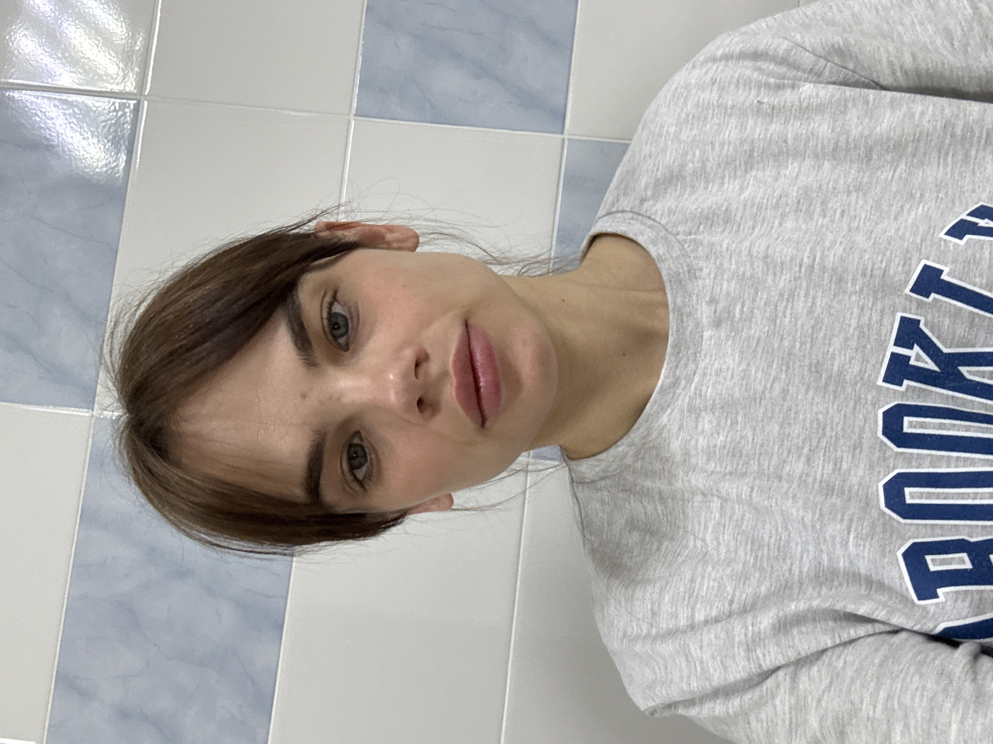 A woman is in what appears to be a bathroom with light blue and white tiled walls. She is wearing a grey sweatshirt with "BROOKLYN" printed on it in blue. Her hair is brown and she has light skin. There is no specific information to indicate the location is in Anapa, Russia.