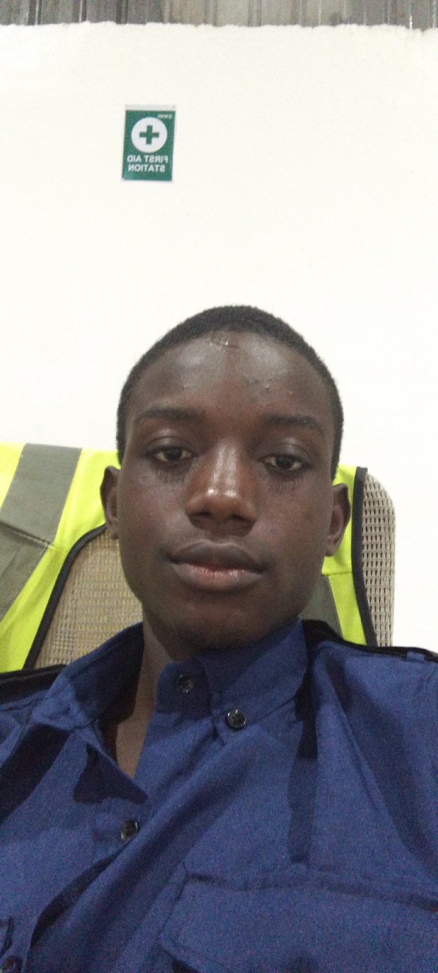 The image shows a man in a blue button-down shirt. Behind him, there is a safety vest draped over the back of a chair, and a "First Aid Station" sign on the wall. The location is Unknown, Unknown.