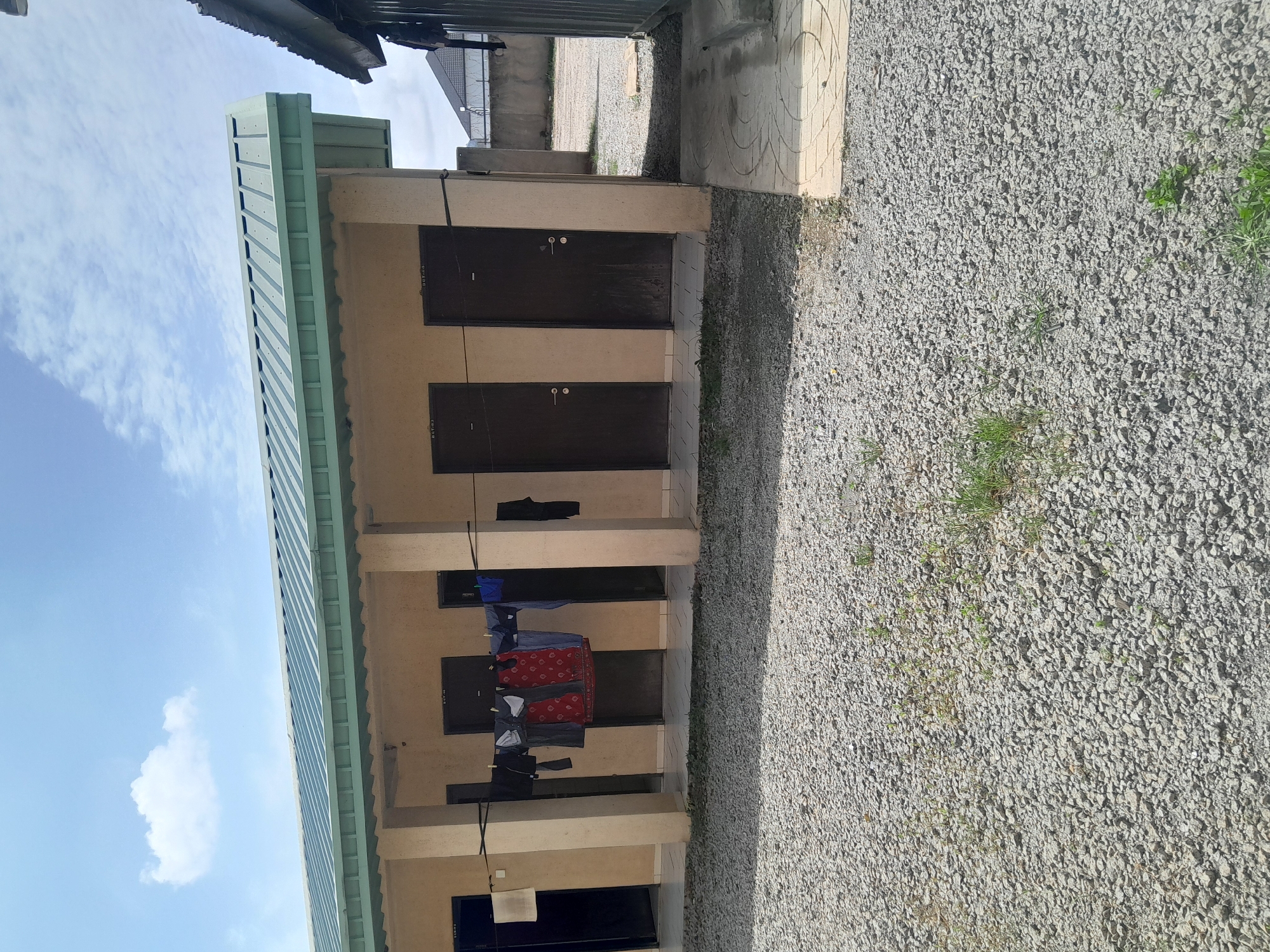 The image shows a row of rooms or units with light beige walls and dark brown doors. A clothesline is strung across the front of some of the units, with clothes hanging on it to dry. The ground in front of the units is covered in small gray gravel, with some patches of green grass. The building has a green, corrugated metal roof. In the background, there is a concrete wall with a fence on top, and a cloudy blue sky. The location is identified as Dam Camp, Nigeria.