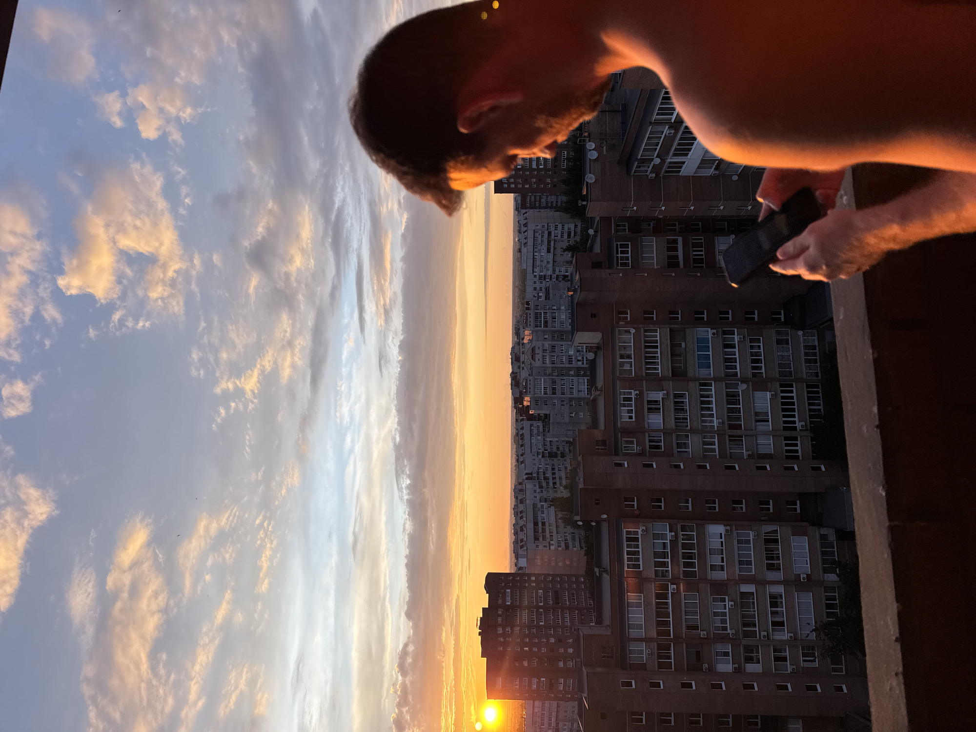The image depicts an urban landscape at sunset, viewed from an elevated vantage point, likely a balcony or rooftop. In the foreground, partially cropped on the right, an adult male, seen from behind and the right side, stands with his back largely facing the viewer. He has short, dark hair and appears shirtless, with his right shoulder and arm prominently illuminated by the warm, orange light of the setting sun. He holds a dark rectangular smartphone with both hands, actively looking down at its screen.
The middle ground is dominated by a dense arrangement of multi-story residential buildings in an unspecified city within "Unknown, Unknown." These buildings feature brick and concrete facades, numerous windows, and visible balconies, some with air conditioning units. A prominent, dark-colored high-rise building stands on the left, partially obscuring the bright, intensely yellow-orange sun, which appears as a double orb due to optical effects.
The background features a vast expanse of sky filled with scattered clouds. The clouds near the horizon are illuminated in vibrant orange, yellow, and pink hues, transitioning to pale blue and grey towards the top of the frame. A faint line on the distant horizon suggests a flat landmass beyond the city. The overall lighting is characteristic of a sunset, casting long shadows and a warm glow across the entire scene. A dark, solid railing or parapet extends across the lower portion of the image, separating the foreground from the urban panorama below.
