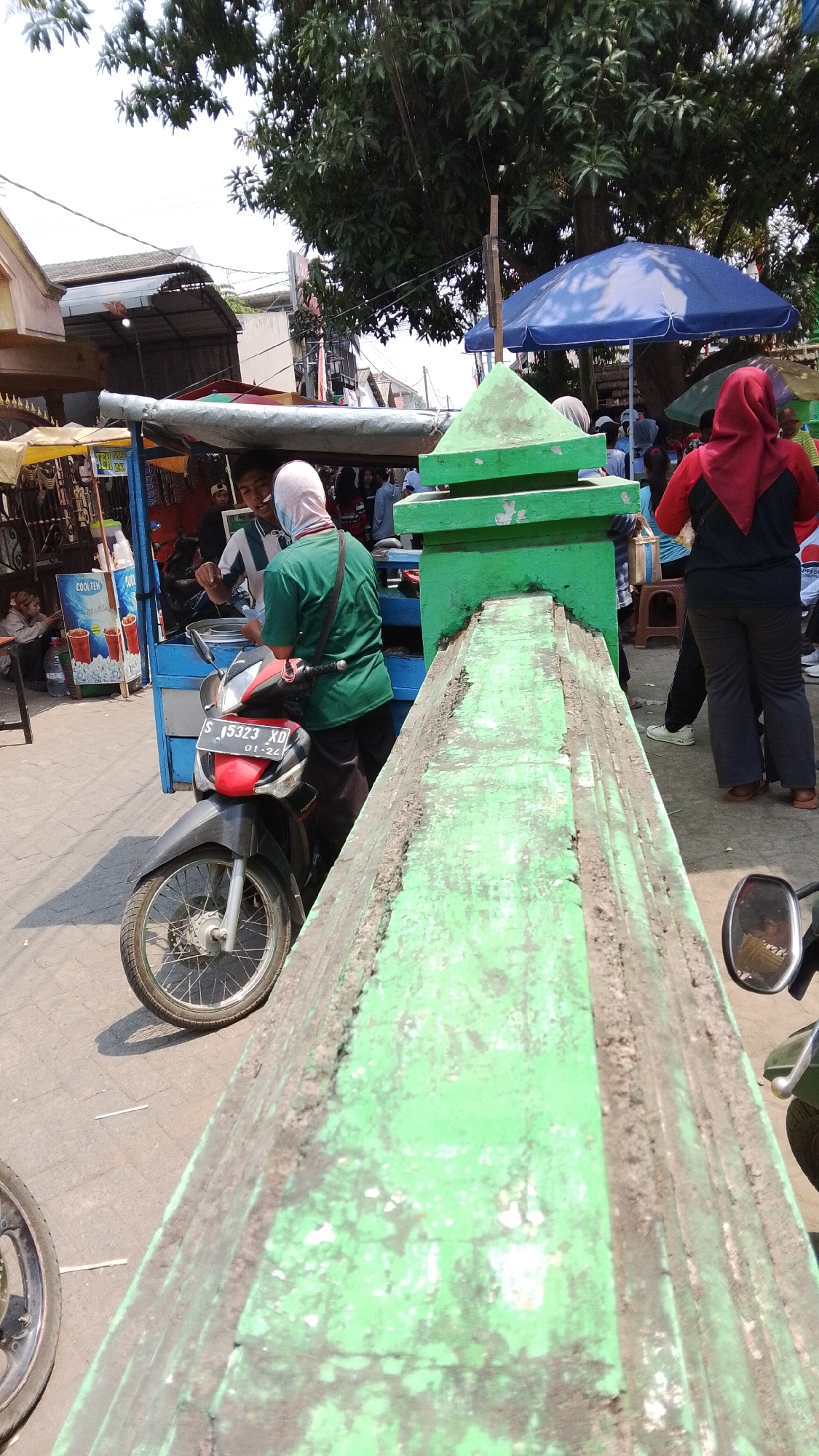 A street scene in Mancar, Indonesia, shows a vibrant marketplace on a sunny day. In the foreground, a person wearing a green shirt and white head covering is on a red and black motorcycle. The motorcycle has a license plate that reads "S 15323 XD 01-24". Behind the motorcycle is a blue cart with a silver awning. To the right of the cart is a bright green, tiered structure. A woman in a red head covering and dark clothing stands to the right of the green structure. In the background, there are more market stalls, trees, and blue umbrellas, indicating a bustling outdoor environment. The ground appears to be paved.