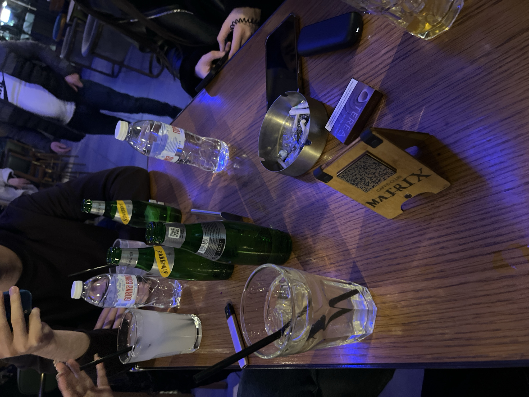 The image captures a dimly lit table scene, seemingly in a bar or cafe. Several people are seated around the table, though they are only partially visible. There are bottles of GORNA BANJA water and Schweppes tonic water. Additionally, there's a glass of clear liquid, possibly a cocktail, with a straw in it. An ashtray filled with cigarette butts, a cell phone, a pack of TEREA cigarettes, and a table stand labeled "MATRIX CAFFE-CLUB" indicate the setting is likely in a social environment. The table is made of wood. The presence of the Matrix Caffe Club suggests that this might be in Goce Delchev, Bulgaria, as it's a likely location for the business.