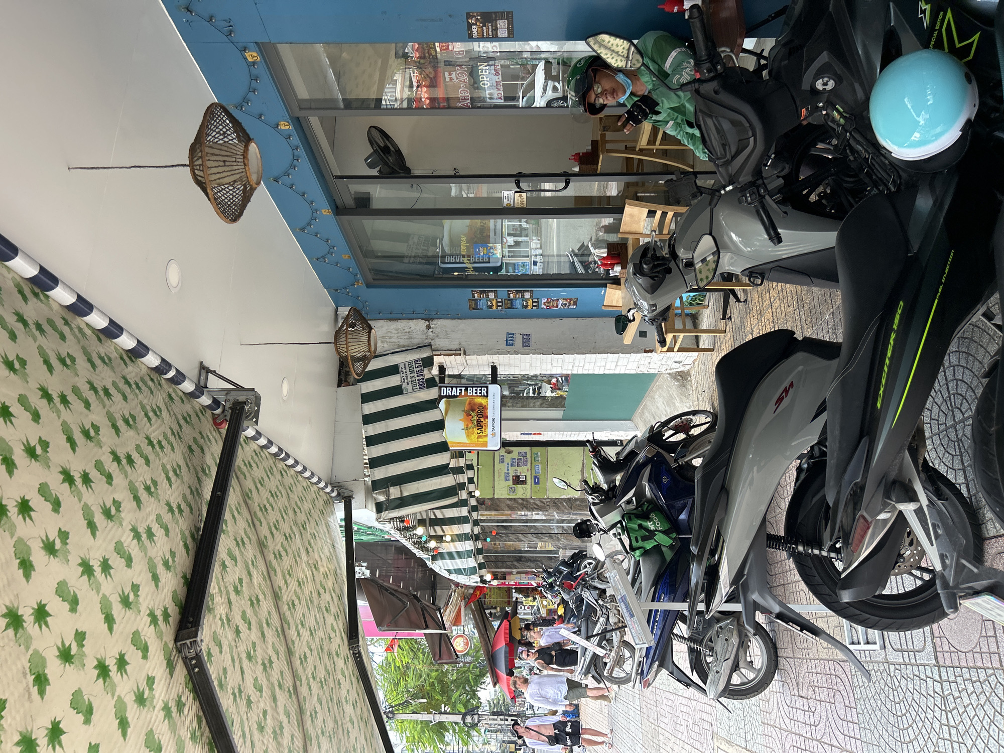 The image, rotated 90 degrees counter-clockwise, captures a bustling street scene in Nha Trang Ward, Vietnam, under bright daylight. The view looks along a narrow street or alley lined with shops and cafes.
Numerous motorbikes are parked closely together on the tiled sidewalk, dominating the right side and foreground of the image. A light blue helmet rests prominently on the handlebars of one motorbike. Another bike has a green bag with "Food" printed on it, suggesting delivery services.
On the left, shopfronts feature large glass windows and doors, some displaying "OPEN" signs. One person, possibly a delivery rider, wearing a green jacket, helmet, and face mask, is visible through a shop window. Inside, tables and chairs are arranged. The building facade above the shops is painted blue and decorated with strings of small lights and woven, basket-like light fixtures. A white awning with a repeating green leaf pattern extends over a portion of the sidewalk, while a green and white striped awning is further down the street.
Several pedestrians are seen walking further down the street, navigating between parked motorbikes and shop fronts. The overall atmosphere is that of an active urban environment.
Visible text includes "DRAFT BEER" and "SAPPORO PREMIUM" on a sign. Other text includes "THIÊN MINH" and the phone number "0933.015.579". "SPECIAL EDITION" is visible on a motorbike. Reflections show distorted text like "OBVEL BEEB" (likely a reversed "DRAFT BEER") and "ОБМЕН" (Russian for 'exchange', potentially a sign or further reflection).