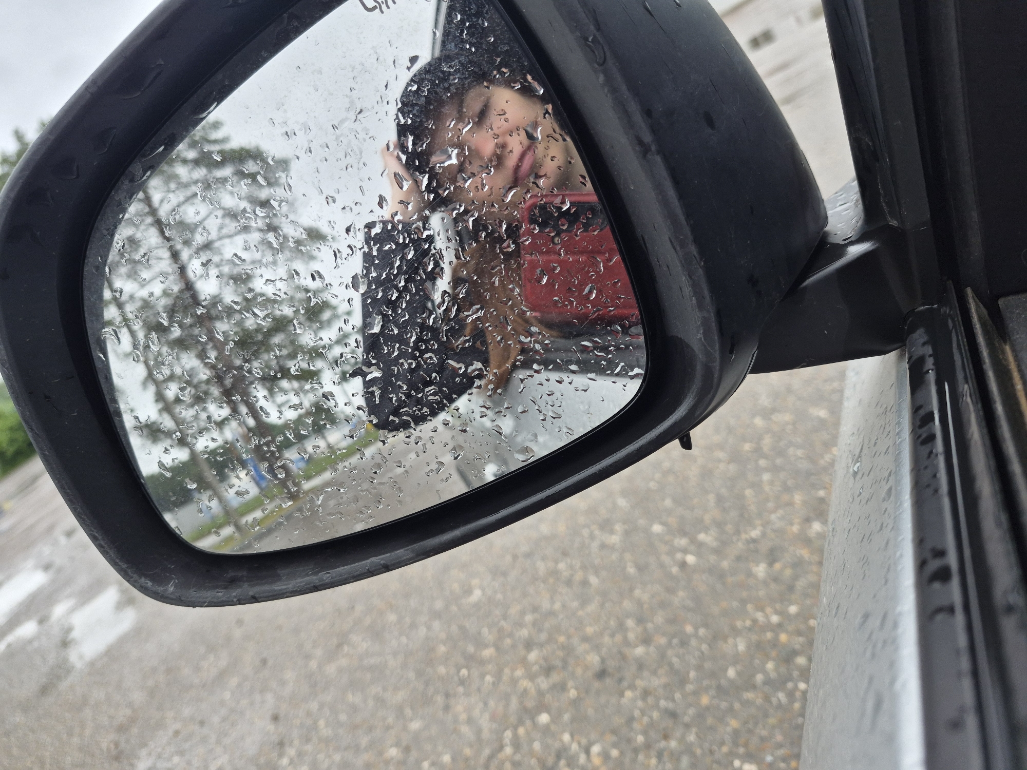 In a side-view mirror of a car, a person is reflected, taking a selfie. The mirror is covered in raindrops, distorting the reflection. The person, with dark hair, is wearing a red shirt and a black hoodie, with their right hand touching the top of their head. A red smartphone is visible in their left hand, used to take the photo. The background in the reflection shows trees and a cloudy sky. The car's exterior is visible in the foreground and left side of the image, with the asphalt road and some white markings also discernible. The weather appears to be rainy.