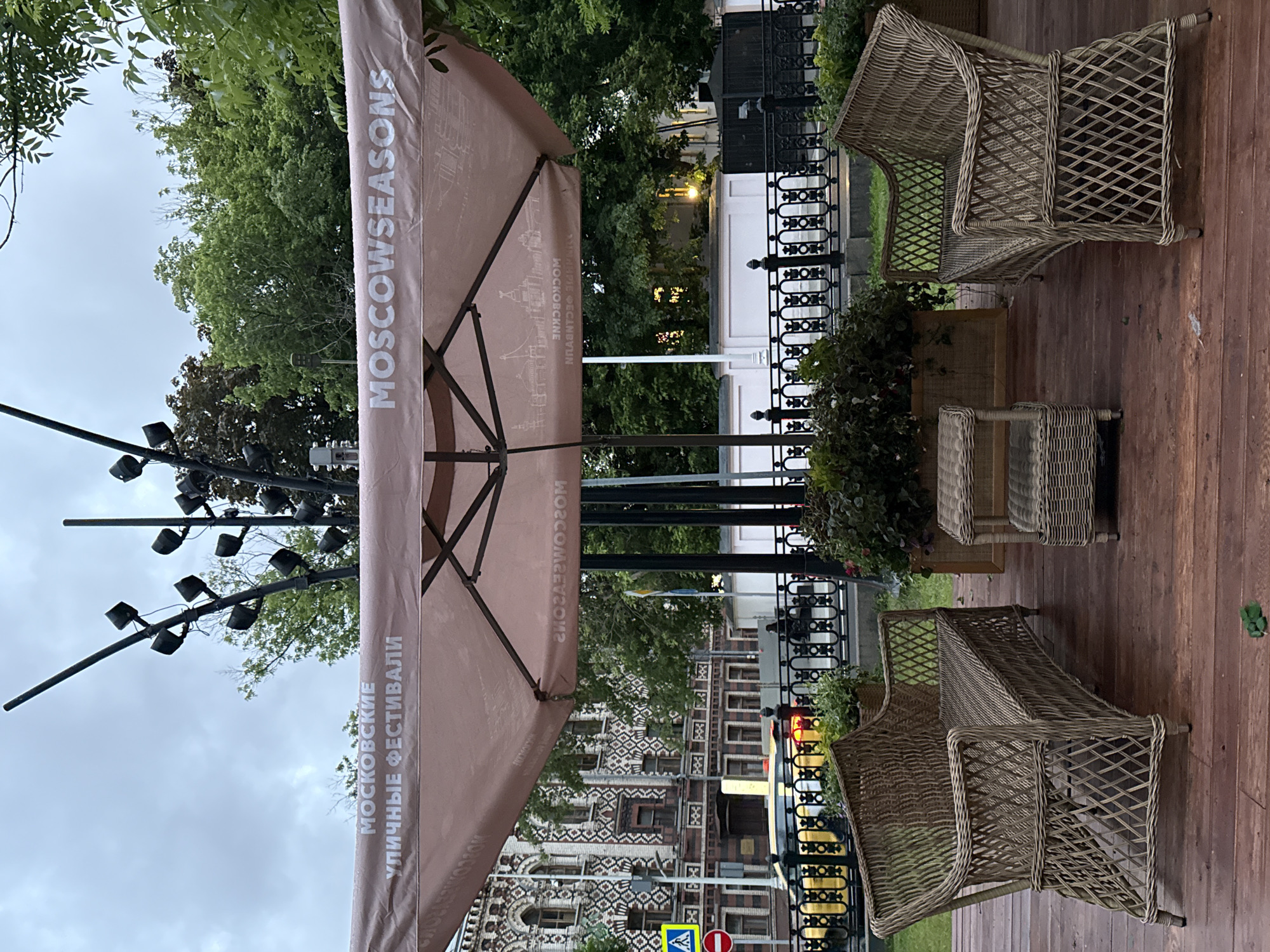 The image depicts an outdoor seating area, likely a café or restaurant patio, situated in Moscow, Russia. The focal point is a large, tilted beige parasol with the text "MOSCOW SEASONS" printed vertically in English and Russian. The Russian text reads "МОСКОВСКИЕ УЛИЧНЫЕ ФЕСТИВАЛИ." Below, the words “Moscow seasons.rf” are written in English. The seating area consists of two wicker chairs and a small wicker table positioned on a wooden deck. A black, ornate metal fence separates the seating area from the street. In the background, a building with patterned brickwork is visible, along with street signs. A light post featuring numerous spotlights is positioned on the left side of the frame. The lighting suggests it is either early morning or evening. The sky appears overcast.