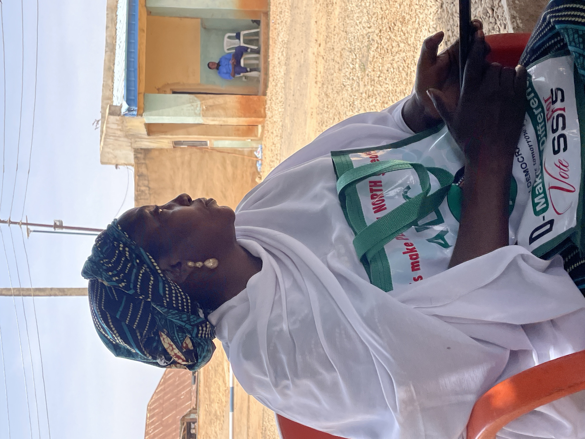 A medium shot depicts an individual seated on a red chair, holding a cell phone. The person wears a patterned head covering and a white garment. A tote bag rests on their lap, displaying political campaign messaging, including the text "Vote SSIS." The background reveals a building with tan walls and a blue awning. In an interior space, a man sits on a white chair. The scene takes place in Bukuru, Nigeria. A utility pole stands in the far left of the image, with multiple wires extending in the sky. The ground appears sandy.