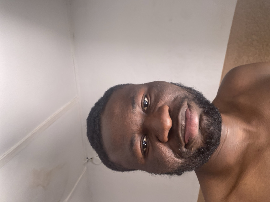 The image displays a close-up portrait of an adult male in an indoor environment located in Udu, Nigeria. The individual, appearing to be of African descent, has a dark skin tone, short dark hair, and a full dark beard and mustache. His dark eyes are directed forward, and his lips are subtly downturned, indicating a neutral facial expression. He is depicted from the shoulders up, appearing shirtless. The background features a predominantly white wall and ceiling. The ceiling shows faint brown discoloration, particularly on the upper left, along with visible linear seams. A small, dark, circular object is affixed to the upper left section of the wall. A horizontal strip of light brown or beige material is visible on the lower right wall. The subject is looking directly at the camera, suggesting a posed photograph or selfie.