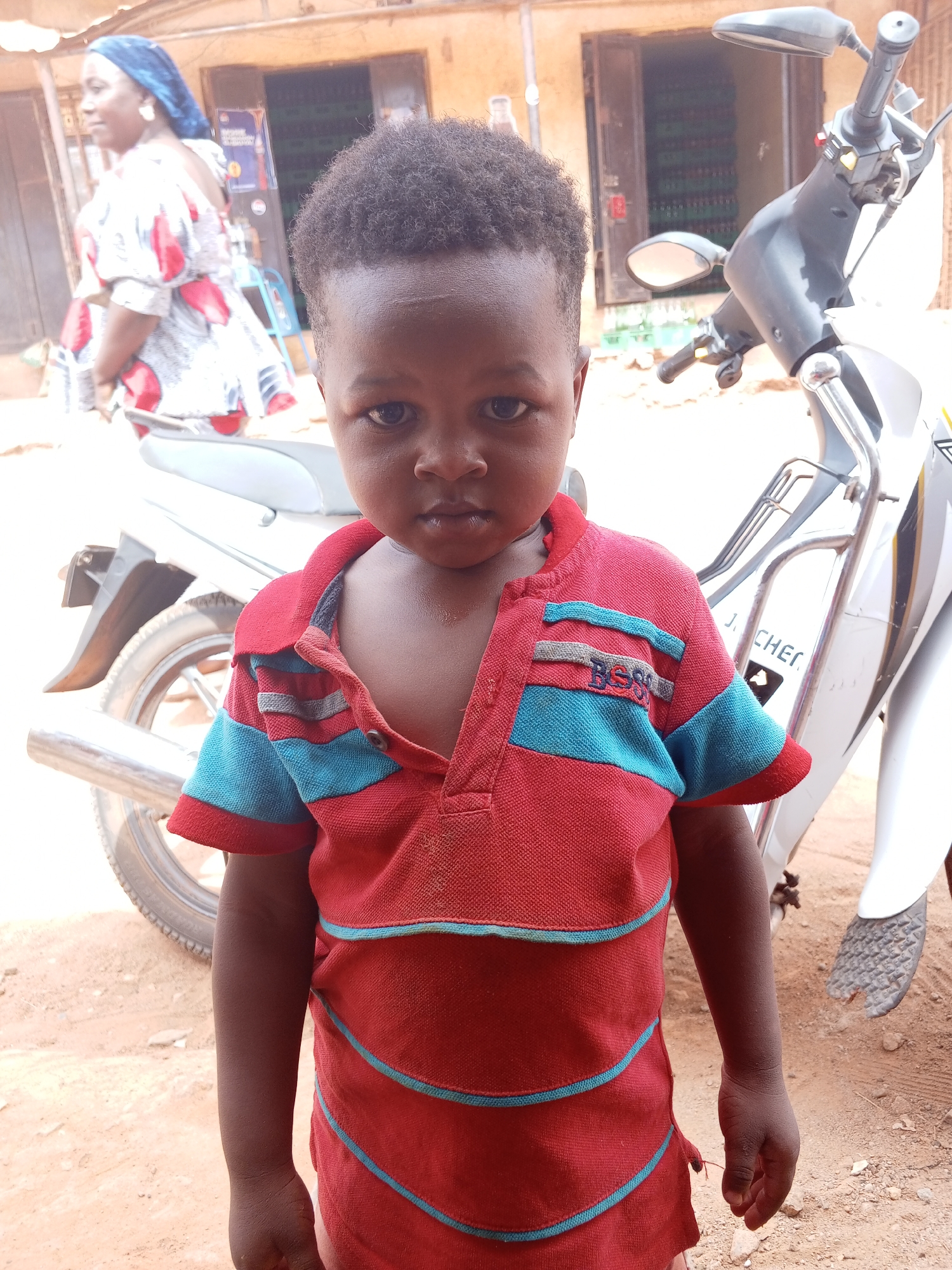 In Yelwa, Nigeria, a young child stands in the foreground, wearing a red and blue striped shirt. Behind him to the right, a white motorcycle is parked. In the background, a woman wearing a blue head covering and a patterned dress walks away from the camera near a building with an open doorway revealing stacked items. The ground is reddish dirt.
