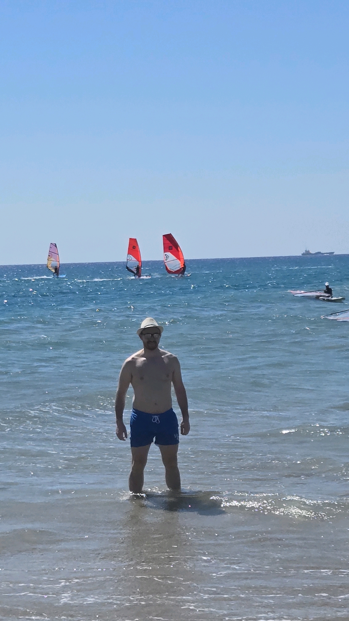 A man wearing blue swim trunks and a straw hat stands in shallow ocean water. He is facing the camera, with windsurfers visible in the background. The sky is clear and blue, and the water is a deep blue. There is a ship visible in the distance. The image appears to have been taken at Prasonisi, Greece.