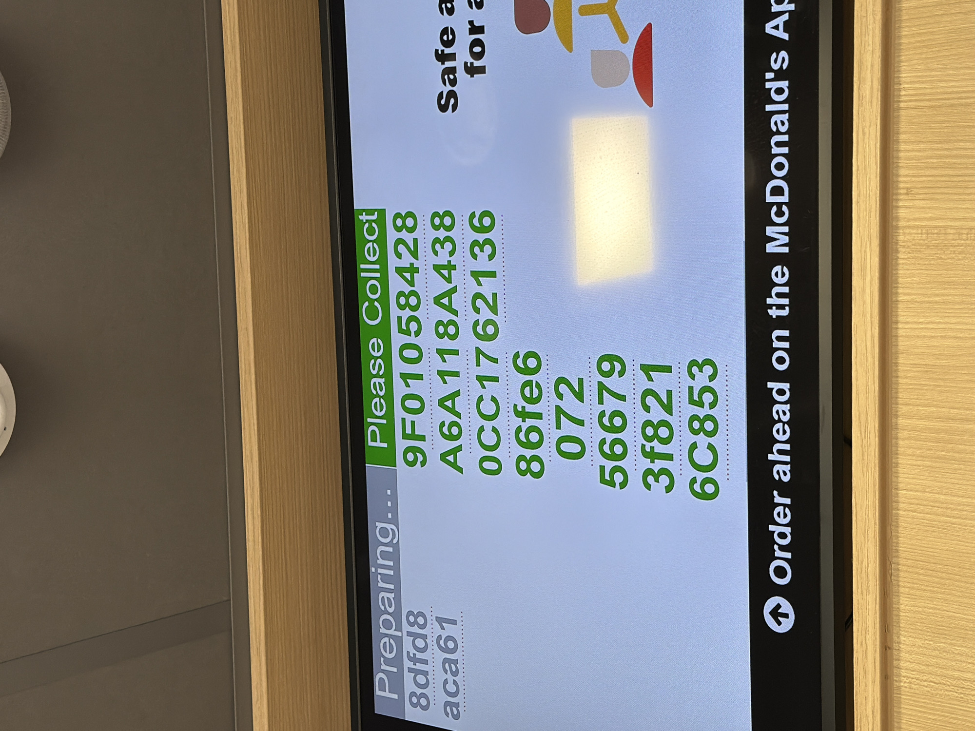 The image shows a digital screen at a McDonald's location, possibly in or near Pity Me, United Kingdom, although the exact location cannot be determined from the image alone. The screen displays order preparation and collection information. On the left, the text "Preparing..." appears, followed by the alphanumeric code "8dfd8 aca61." In the center, under the heading "Please Collect," a list of alphanumeric order codes (9F01058428, A6A118A438, 0CC1762136, 86fe6, 072, 56679, 3f821, 6C853) is displayed. To the right of the order codes, there's a visual element suggesting a safe and the wording "Safe a for a". At the bottom, an arrow icon precedes the text "Order ahead on the McDonald's Ap." The screen is framed by a wooden surface.