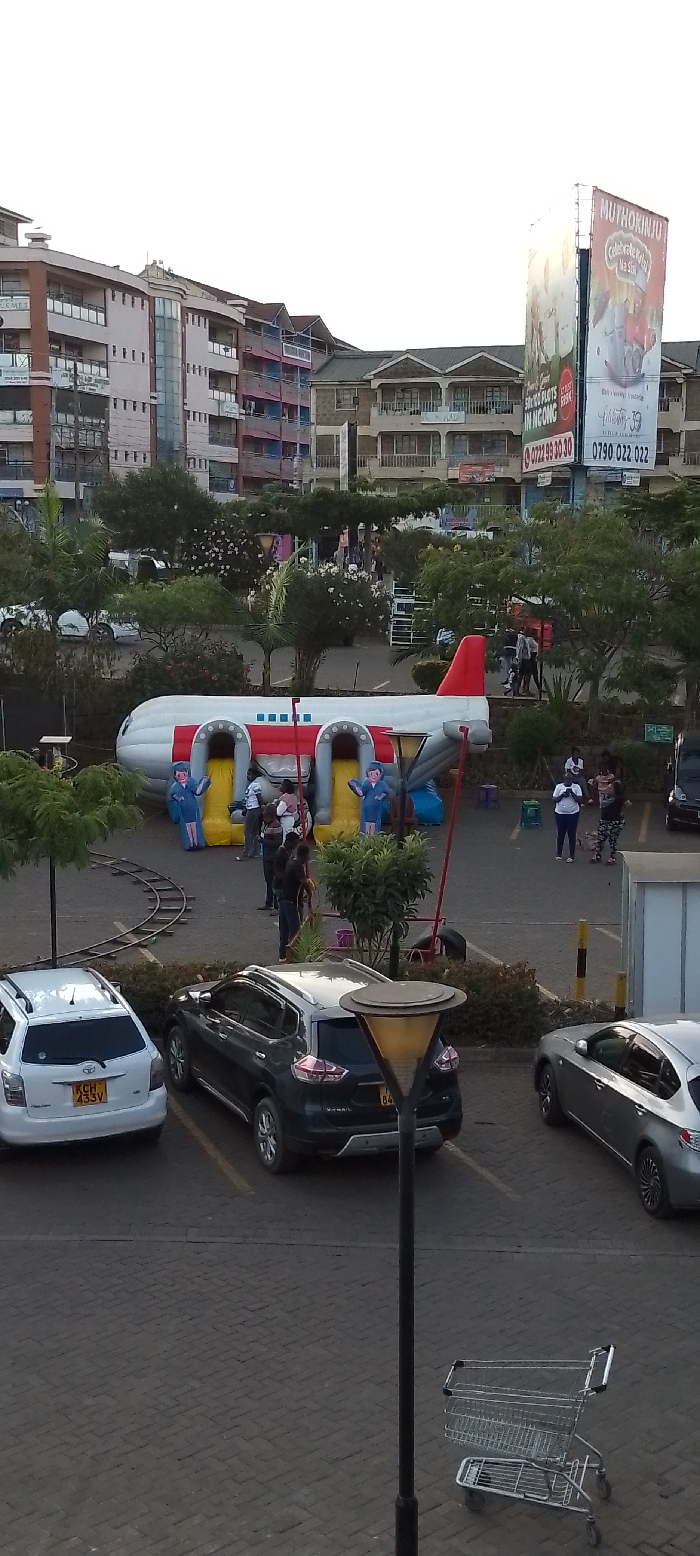 The image depicts an outdoor urban environment located in Ngong ward, Kenya. A paved central area features an inflatable bounce house designed as a white and red passenger airplane, equipped with two yellow slides and two inflatable figures resembling airline staff at the base of the slides. Approximately eight to ten individuals, comprising both adults and children, are present around and within the inflatable structure, engaged in recreational activity. Sections of a miniature train track are visible on the paved ground to the left of the inflatable.
In the foreground, a paved parking lot contains three parked vehicles: a white Toyota SUV/wagon (license plate KCH 433V), a dark grey SUV (partial license plate K*** 84), and a silver sedan. A black street lamp post and a metal shopping cart are positioned in the lower foreground. The background is dominated by multi-story buildings, exhibiting various architectural styles and colors, indicative of residential or commercial use. A large, illuminated billboard on the right side of the background displays advertisements, including the text "MUTHOKINJU," contact numbers "0722 99 30 30" and "0790 022 022," and "50x100 PLOTS IN NGONG," providing specific location context. Trees and shrubs are interspersed throughout the scene, separating structures and parking areas. The sky is clear, indicating daylight conditions.