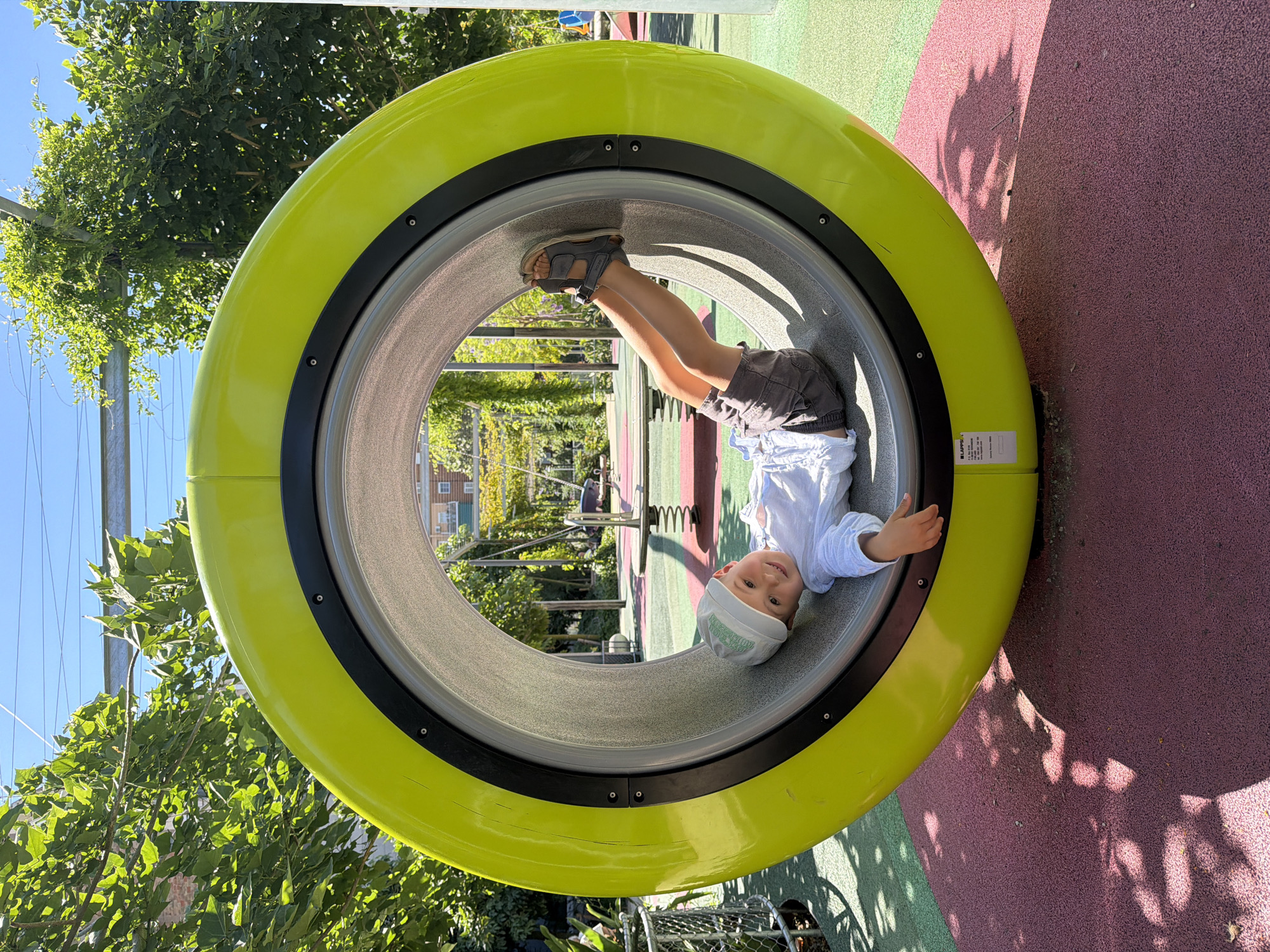 A young boy is lying on his back inside a large, yellow, circular tunnel. He is wearing a white long-sleeved shirt, grey shorts, a light-colored baseball cap, and sandals. His head is tilted to his right, and he is looking at the camera with a smile. His legs are bent, with his right foot resting on the inside of the tunnel. The tunnel is part of a playground. The ground is a multi-colored soft surface, with areas of pink and green. In the background, there are trees with green leaves, a blue sky, and playground equipment, including a spring rider. The scene appears to be in a park or public space. The location is identified as Parc del Fòrum in Barcelona, Spain.
