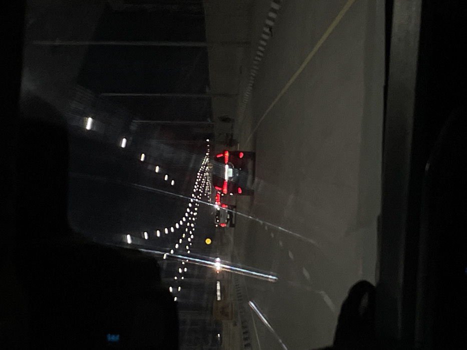 The image depicts a nighttime scene from the interior of a moving vehicle on a multi-lane roadway in Alaguntan, Nigeria. The perspective is through the front windshield, exhibiting reflections from internal dashboard elements and external streetlights. Ahead, three vehicles are visible; the closest is a dark sport utility vehicle (SUV) with illuminated red tail lights, followed by a lighter-colored pickup truck, and another vehicle further in the distance. The road is asphalt, marked with white dashed lane lines and a solid yellow line on the right shoulder. Overhead, numerous streetlights arranged in an arc illuminate the thoroughfare. Black and white striped concrete barriers delineate the left and right sides of the road. On the far right, alongside the barrier and a dark wall, a lone, indistinct dark figure, possibly a person, is visible. The environment is dark, suggesting a clear night with artificial illumination as the primary light source.