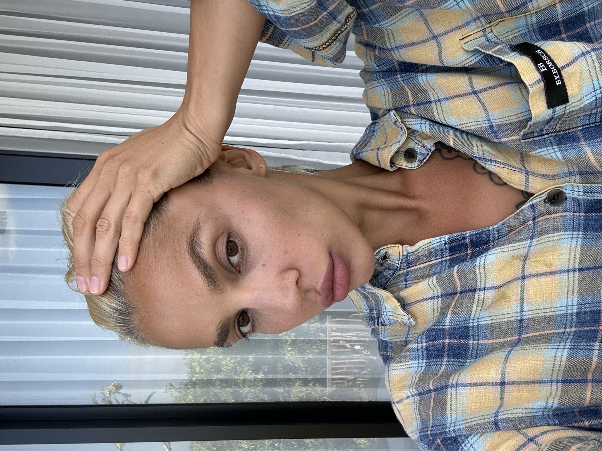 A woman with blonde hair and visible dark roots, brown eyes, and full lips is depicted in a close-up, head-and-shoulders shot, looking directly into the camera. Her right hand is placed on the crown of her head, with fingers spread across her hair. She is wearing a blue, yellow, and light pink plaid long-sleeved button-up shirt, partially unbuttoned to reveal a portion of her upper chest and a dark-colored tattoo. A black label with white text "BB BY.BORSCH" is affixed to the left chest pocket of her shirt. The background features a window or glass door with light-colored vertical blinds or panels creating distinct vertical lines. Beyond the window, green foliage and a dark-colored railing structure are visible. The scene is located in Kuta Selatan, Indonesia.