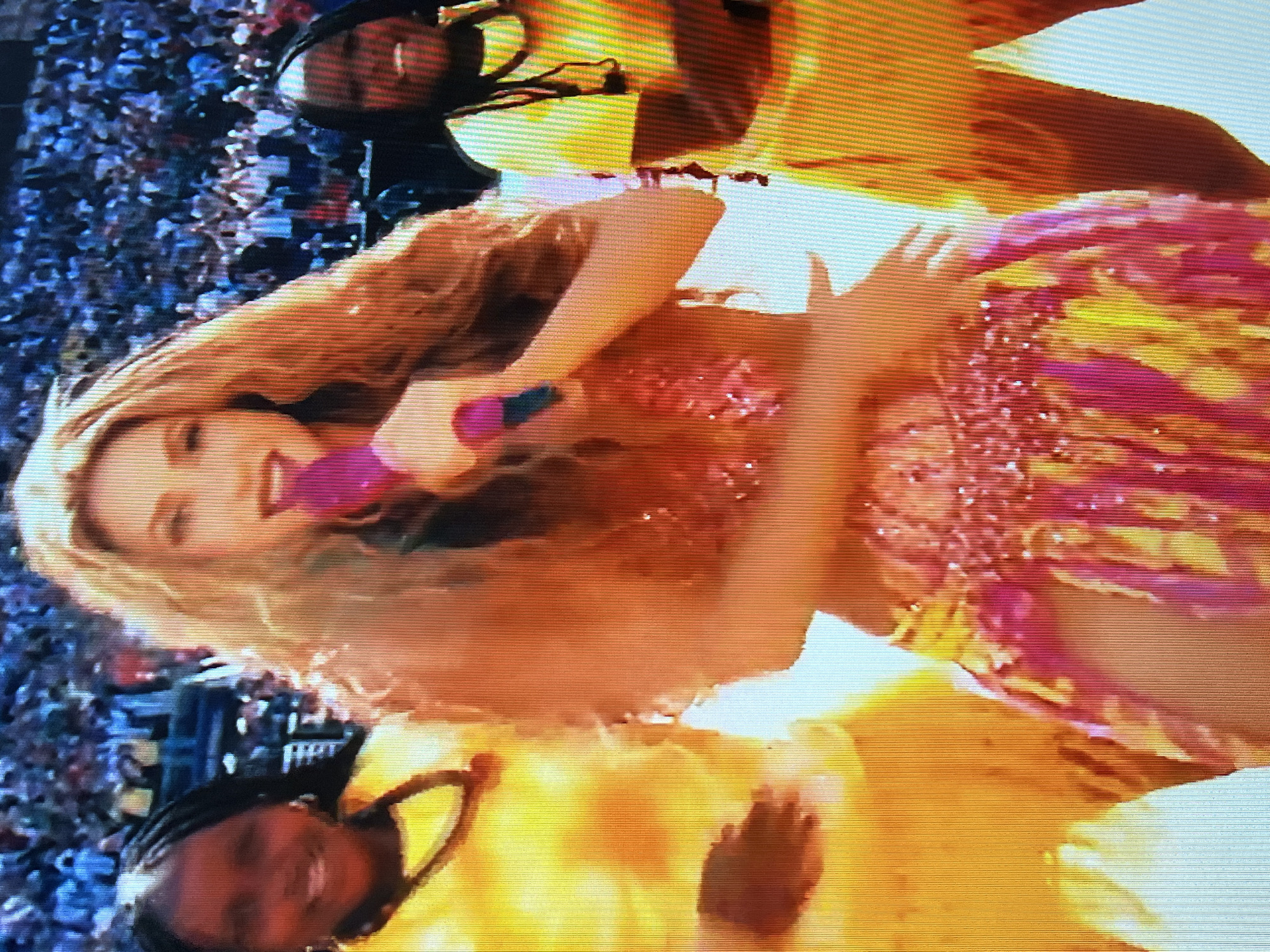 The image captures a vibrant performance with a central figure, Shakira, singing into a microphone. She is adorned in a sequined, colorful costume featuring shades of pink, yellow, and red, with a flowing skirt. Flanking her are two young girls, dressed in bright yellow outfits and also appearing to be performing or dancing. The background is blurred but suggests a large crowd of spectators, indicating an outdoor event or stadium setting. The overall atmosphere appears energetic and festive. The image does not provide any information to locate the scene in Moscow, Russia, or any other specific geographical location.