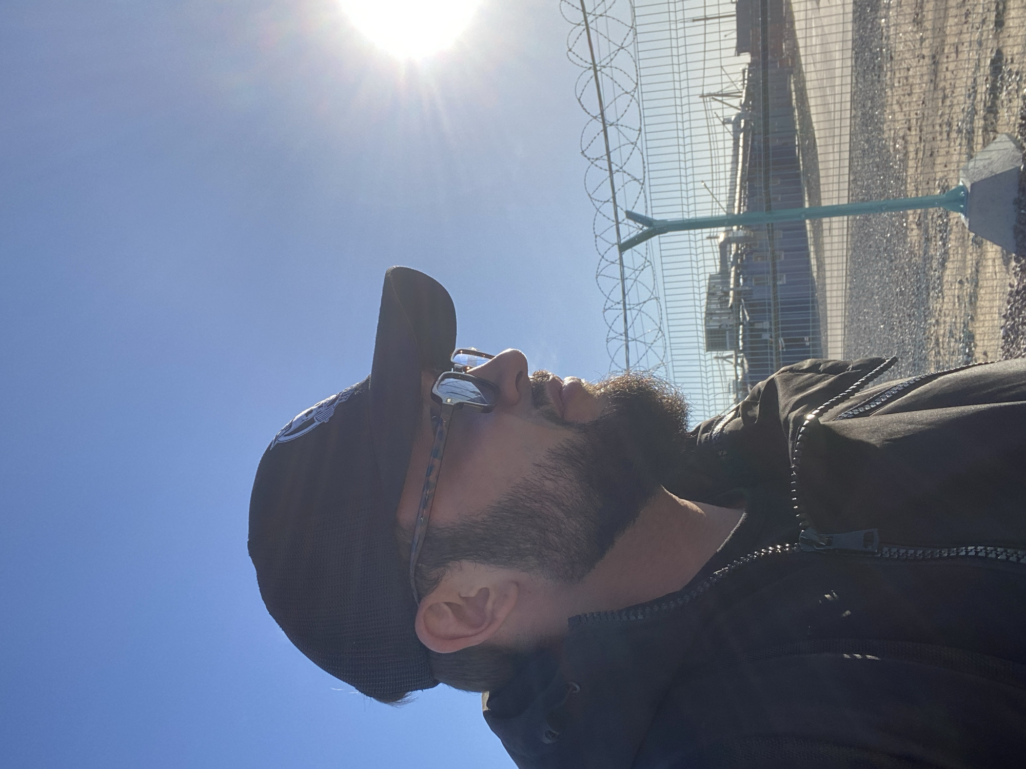The image shows a man wearing a black baseball cap and sunglasses, looking to his right. He has a beard and is wearing a black jacket. Behind him, a barbed wire fence separates him from a background that appears to be an industrial area with shipping containers and buildings. The sky is bright blue with the sun shining, creating a lens flare on the right side of the image. The location is stated to be the city of Koskolovo, Russia.