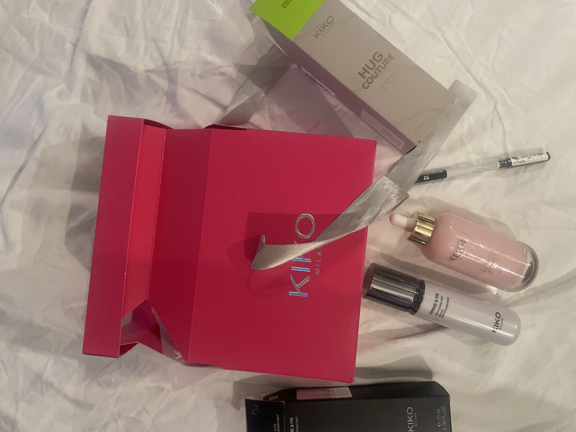 An overhead view depicts an arrangement of Kiko Milano cosmetic products laid out on a white, wrinkled fabric surface. Central to the scene is a large, open, fuchsia-colored Kiko Milano box featuring the brand name in silver, reflective lettering. A silver-grey satin ribbon is draped over this box, extending towards the lower right. To the left, a black rectangular Kiko Milano box, labeled "PRIME & FIX REFRESHING MIST" and indicating a volume of "e 70 ml 2.36 FL.OZ.", stands upright. Below this box, a white spray bottle with a silver cap, also marked "KIKO MILANO PRIME & FIX REFRESHING MIST," rests horizontally. Adjacent to it, a clear glass dropper bottle with a gold cap, containing a light pink liquid and labeled "KIKO MILANO HUG COUTURE," is positioned. To the right of the fuchsia box, a light grey rectangular Kiko Milano box with a neon green top stripe, labeled "HUG COUTURE," lies horizontally. Partially obscured by the ribbon, a slender black and clear makeup pencil, marked with the number "33," is visible. The scene is illuminated by artificial light, creating soft shadows. This arrangement is located in Sofia, Bulgaria.