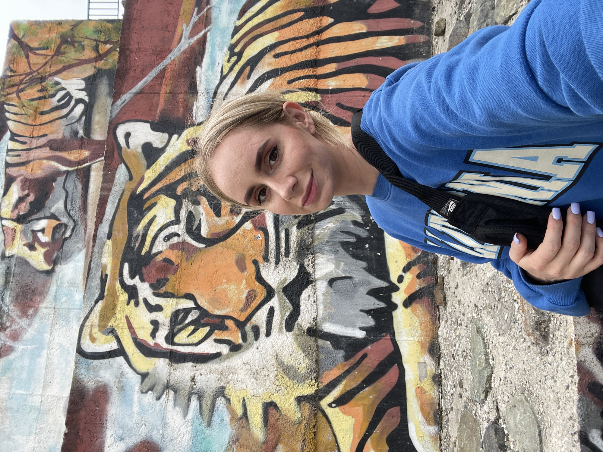 A young woman stands facing the camera, angled slightly to the left, posing in front of a concrete wall adorned with a mural depicting a tiger. The wall is textured and rough, showing signs of weathering and age. The tiger mural is painted in shades of orange, yellow, black, and white, with thick outlines. The woman is wearing a light blue sweatshirt with white lettering. A black bag is slung across her body. Her hair is blonde, styled back from her face. Her fingernails are painted a light purple color. The scene is an outdoor urban setting, possibly a public space within the city of Vladivostok, Russia, given the presence of the tiger mural which is a common motif for this city.