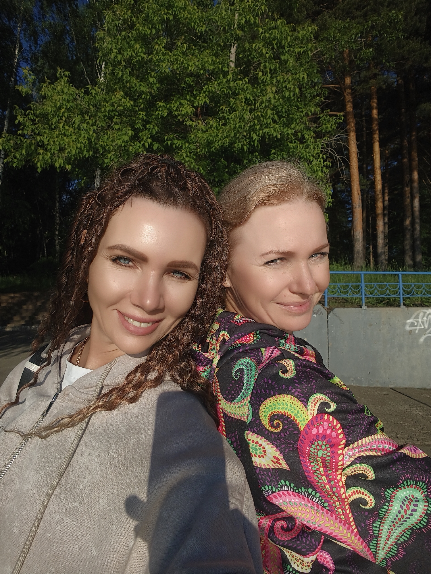Two women are captured in a bright outdoor portrait, leaning back-to-back or side-by-side and smiling. The woman on the left has long, dark brown hair styled in tight curls or micro-braids, and piercing blue eyes. She is smiling broadly towards the camera, wearing a light beige hooded sweatshirt over a white t-shirt, with a gold chain visible around her neck. A distinct shadow falls across her right shoulder and arm.
The woman on the right has blonde hair and is looking over her left shoulder towards the camera with a subtle smile. She is wearing a dark-colored top adorned with a vibrant, intricate pattern of swirling, teardrop, and dotted motifs in shades of pink, green, yellow, and purple.
The background features lush green trees, with lighter-foliaged trees on the left and taller, darker pine trees on the right. Below the trees, in the mid-ground, there is a low concrete wall or structure with a blue metal railing on top. Some indistinct graffiti or tagging is visible on the concrete surface behind the right woman.
The scene is brightly lit by strong sunlight, indicating either late morning or late afternoon, creating clear highlights on the subjects' faces and hair, and casting defined shadows. The overall mood is cheerful and relaxed. The setting appears to be a park or a natural area adjacent to a public walkway or embankment.