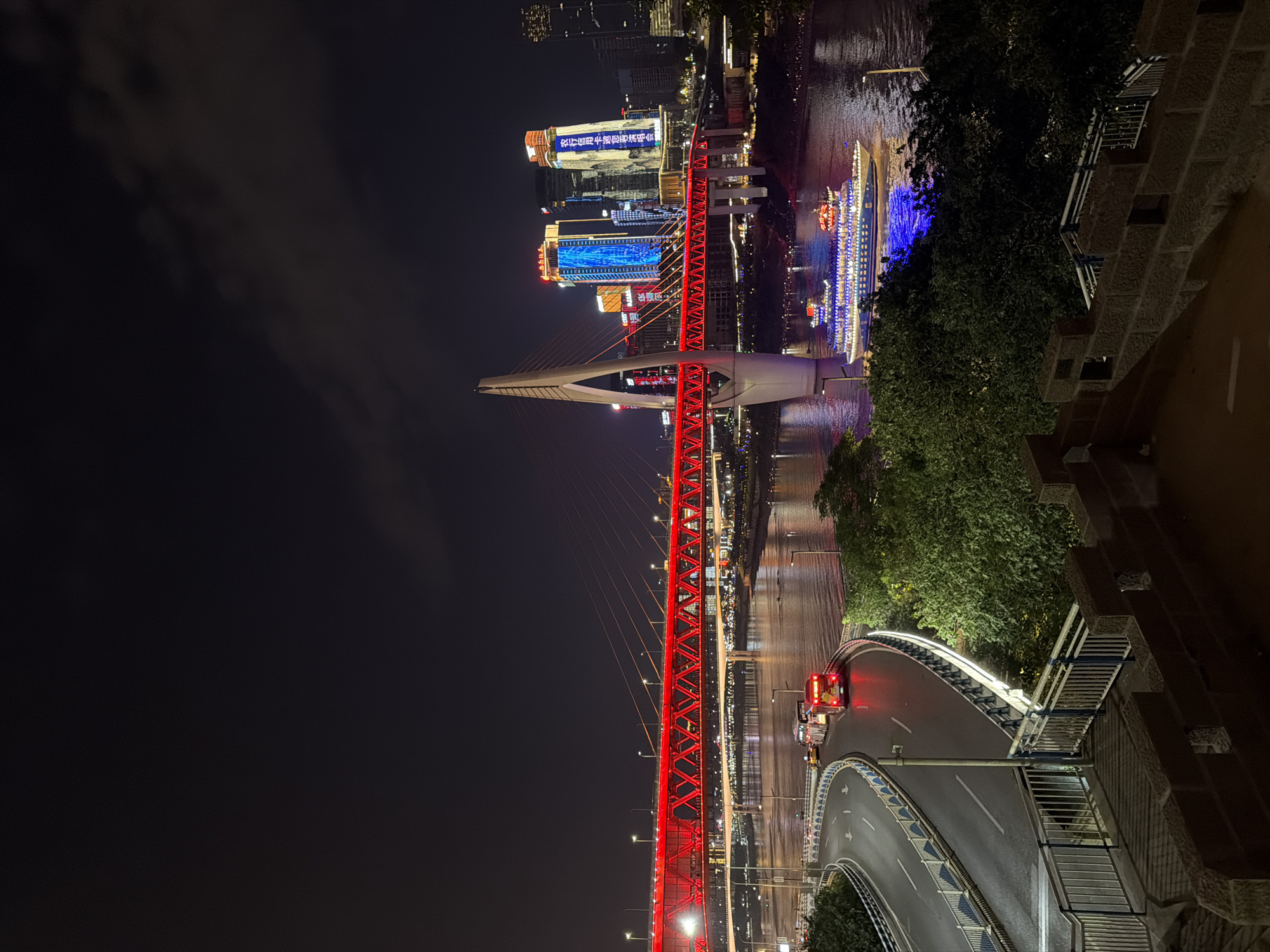 The image depicts a nighttime cityscape in Yuzhong District, China, featuring the iconic Hongyadong area and the Yangtze River. A prominent red suspension bridge, illuminated with bright red lights, spans across the river. To the right of the bridge, modern skyscrapers with vibrant, illuminated facades rise into the dark sky, some displaying Chinese characters. In the foreground, a curved road with white lane markings leads away from the viewer, with several vehicles, including a red bus with its brake lights illuminated, present on the road. A body of water, the Yangtze River, flows beneath the bridge, reflecting the city lights. A large, multi-decked boat, decorated with numerous blue and white lights, is cruising on the river. Lush green trees are visible on the riverbanks and in the foreground, adding a natural element to the urban scene. The overall atmosphere is one of a bustling, illuminated metropolis at night.