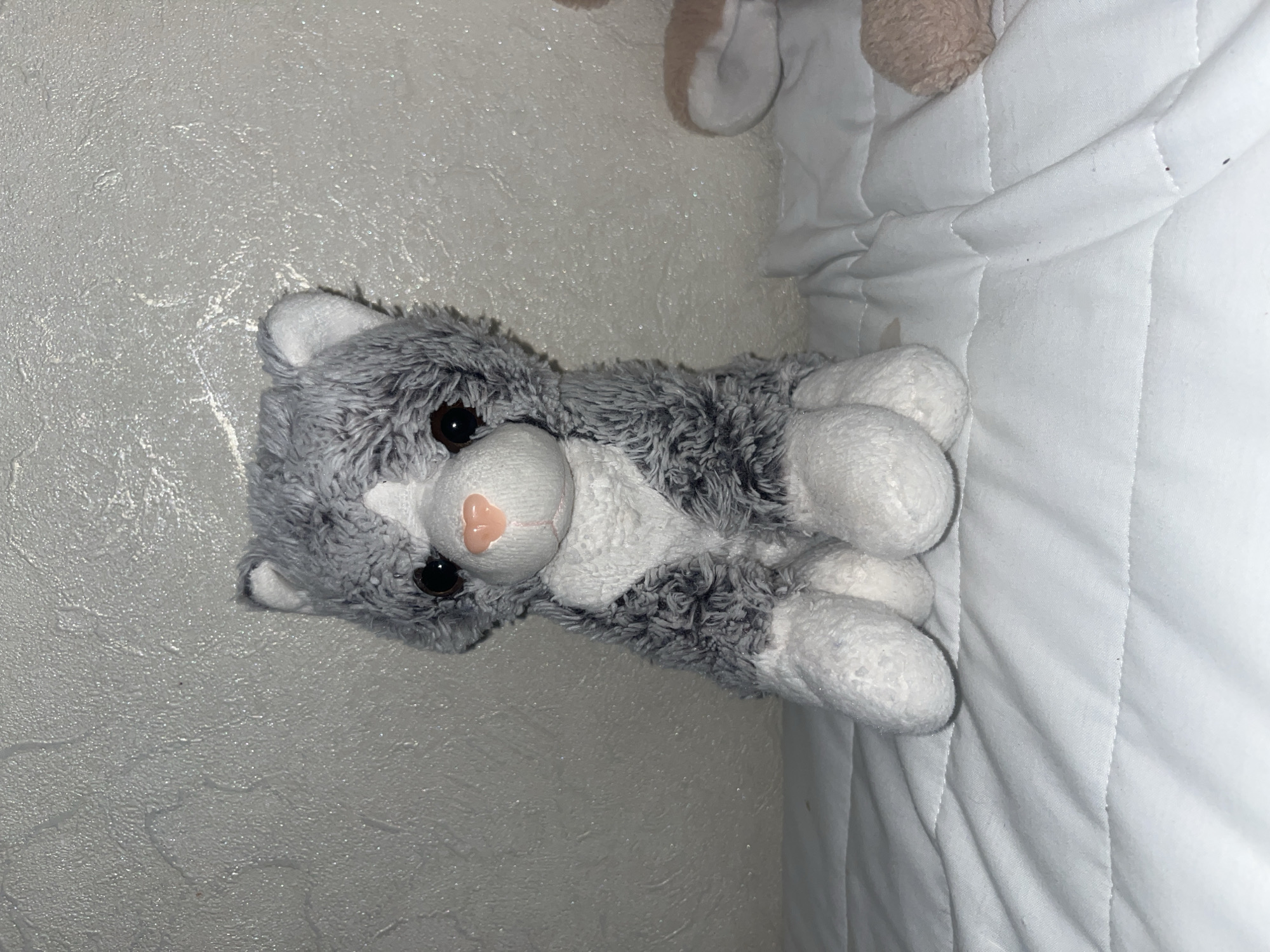 A plush toy cat sits on a white quilted surface. The cat is gray and white with black eyes and a pink nose. The background is a textured, light gray wall with some subtle white markings. To the right of the cat, a portion of another plush toy, possibly a bear, is visible. The lighting appears to be artificial and casts shadows. The scene is indoors and appears to be a bedroom or similar domestic setting, though the specific location within the city of Unknown, Unknown cannot be determined from the image alone.