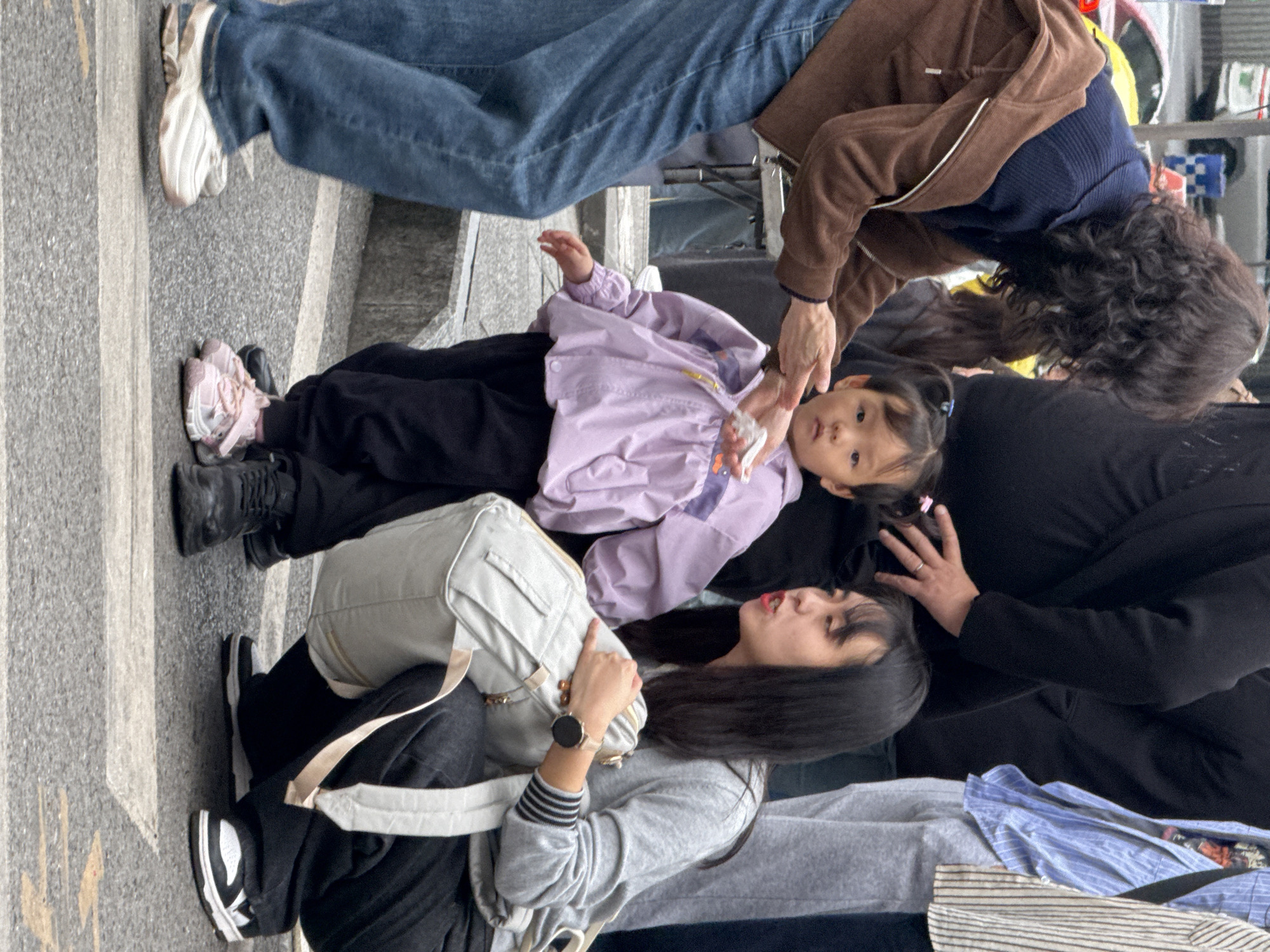 This image captures an urban street scene in Yuzhong District, China, involving three primary individuals and several background figures.
In the foreground, a young child, appearing to be approximately 2-4 years old, stands centrally. The child wears a light purple long-sleeved jacket with a ruffled bottom, black pants, and pink athletic shoes with white soles and hook-and-loop fasteners. The child has dark hair styled in two small pigtails and looks directly towards the viewer with a neutral expression.
To the child's left, partially obscured, is an adult female with dark, curly hair. She wears a dark blue top and a brown zip-up jacket over blue jeans. Her hands are extended towards the child, palms open and slightly cupped, appearing to present or receive a small, crumpled white object held in the child's right hand. Her posture suggests she is leaning down towards the child.
To the child's right, an adult female is crouched down to the child's eye level. She has long, straight dark hair and wears a grey long-sleeved top with black and white striped cuffs, black pants, and black and white athletic shoes. She is holding a light grey backpack with tan straps, resting it on her lap while crouching. Her mouth is open as if speaking, and her gaze is directed towards the child. A dark-faced smartwatch is visible on her left wrist.
Several other individuals are partially visible in the background, out of focus. One person in a black top stands behind the curly-haired woman, and another in a light blue striped shirt and grey sweatpants is behind the crouching woman. The setting appears to be a paved street or sidewalk, with a concrete curb or steps visible behind the child and a blurry vehicle in the distant background. The ground is a speckled grey pavement, with a white line marking a pedestrian crossing or boundary.