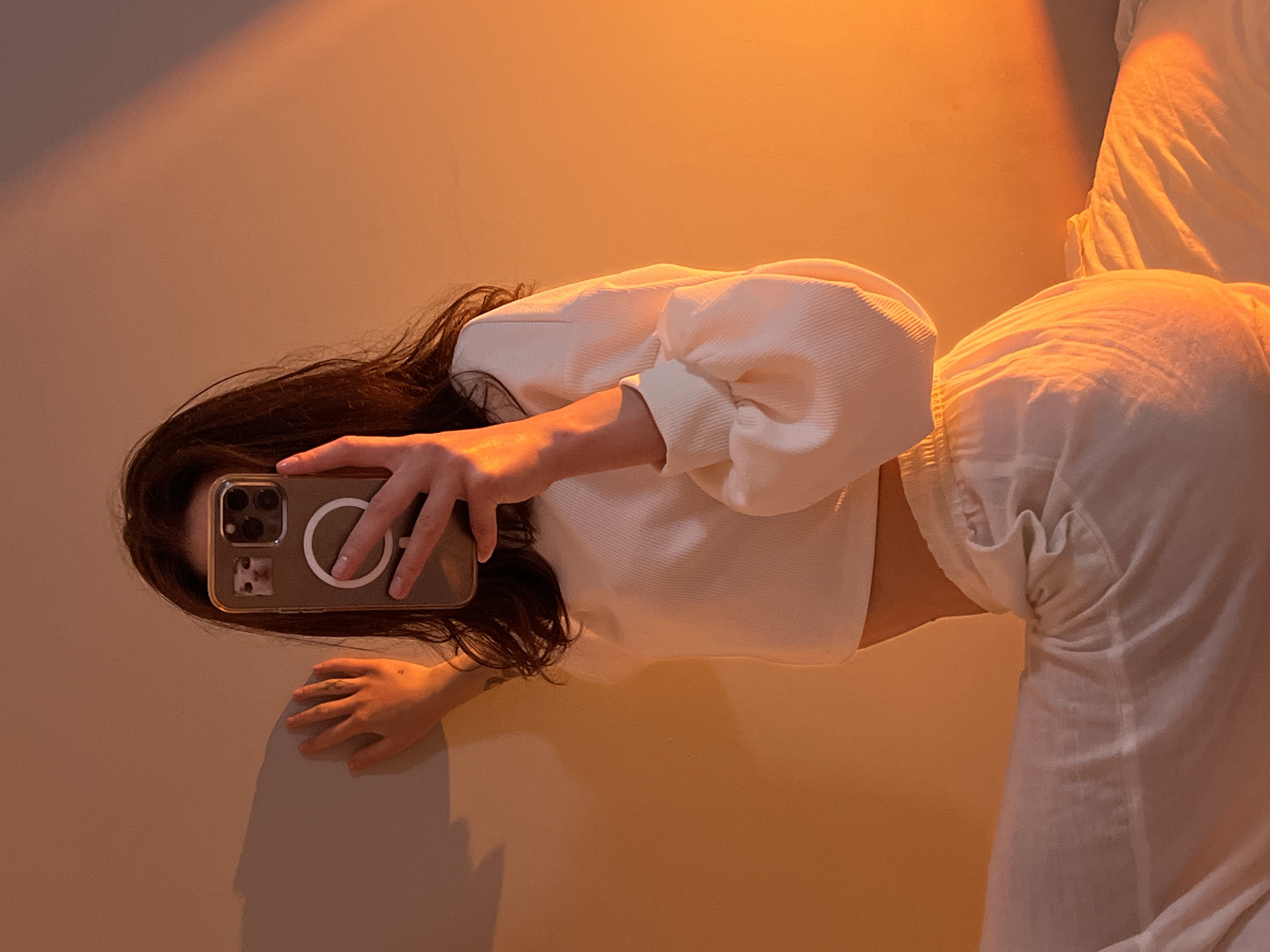 A woman is taking a mirror selfie in a room. She is wearing a white crop top with voluminous sleeves and white pants. She is holding a phone with a case that has a picture of a cat on it. The room is lit with warm, orange light, creating a dramatic shadow on the wall behind her. The scene appears to be indoors, possibly a bedroom, with a white bed visible. There is no information to suggest the location is Ternopil, Ukraine.