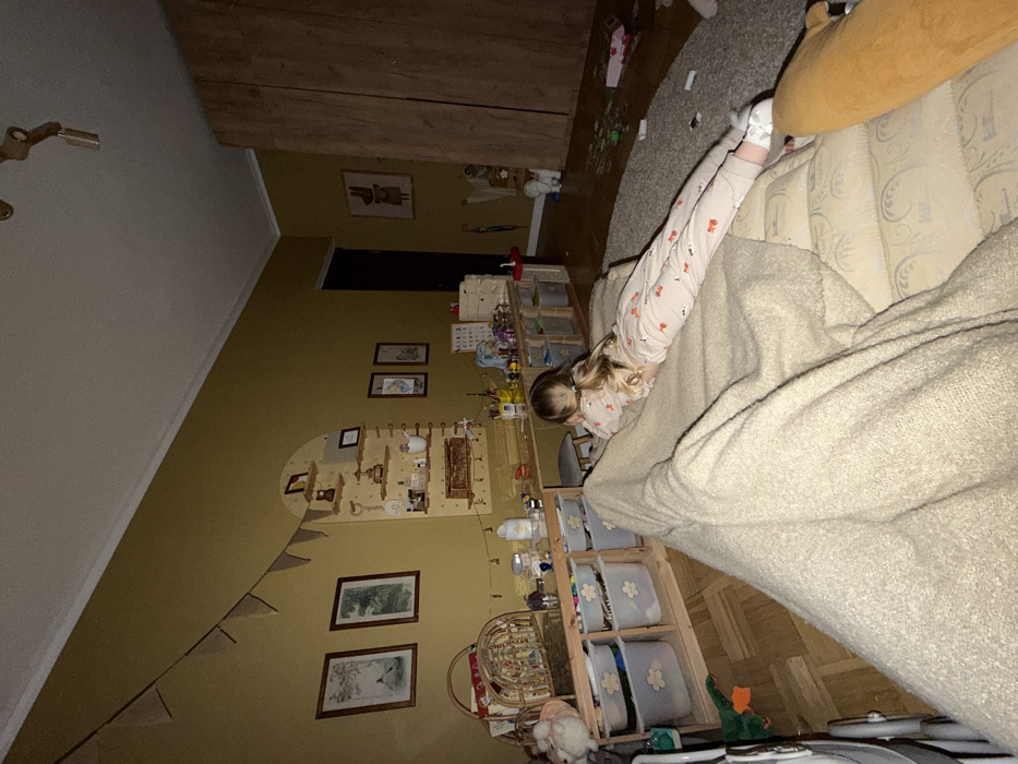 This image depicts an indoor domestic scene in Искра Снт (чашниково), Russia, characterized by subdued artificial lighting. The room's walls are painted a uniform mustard yellow, contrasting with a white ceiling and crown molding. The floor is covered with light-colored wood planks in a herringbone pattern on the left and a light grey textured rug on the right.
A young child, appearing female with long blonde hair tied in a ponytail, is the sole visible individual. She is lying prone on a light-beige upholstered sofa or bed in the foreground, facing away from the camera, and looking towards the far wall. She is dressed in light-colored pajamas adorned with a repeating pattern, possibly foxes, and white socks or slippers. A beige textured blanket is draped over the sofa, partially covering her.
Along the main wall, an arched wooden shelving unit holds various decorative items and small bottles. Below it, a long, low wooden desk or table extends, featuring numerous art supplies, toys, and miscellaneous objects on its surface. Underneath the desk, multiple light-grey plastic storage bins with flower-shaped cutouts are organized within wooden cubbies. A small grey-cushioned stool is tucked beneath the desk. To the left of this setup, two framed botanical prints are hung on the wall. A string of small, triangular pennants hangs diagonally from the upper left corner of the room towards the arched shelving.
To the right, a dark vertical opening, likely a doorway, is visible. Adjacent to it stands a tall, light-colored wooden wardrobe with two panel doors. Between the doorway and the wardrobe, a framed print of a stylized rabbit or animal is displayed on the wall. On the floor, near the wardrobe and on the rug, various items are scattered, including a white stuffed animal, a small red umbrella, and several indistinct objects, with what appears to be puzzle pieces on the rug. A green toy, possibly a duck or dinosaur, rests on the wooden floor in the midground. A brown/orange stuffed animal or cushion is partially visible on the far right of the sofa in the immediate foreground.