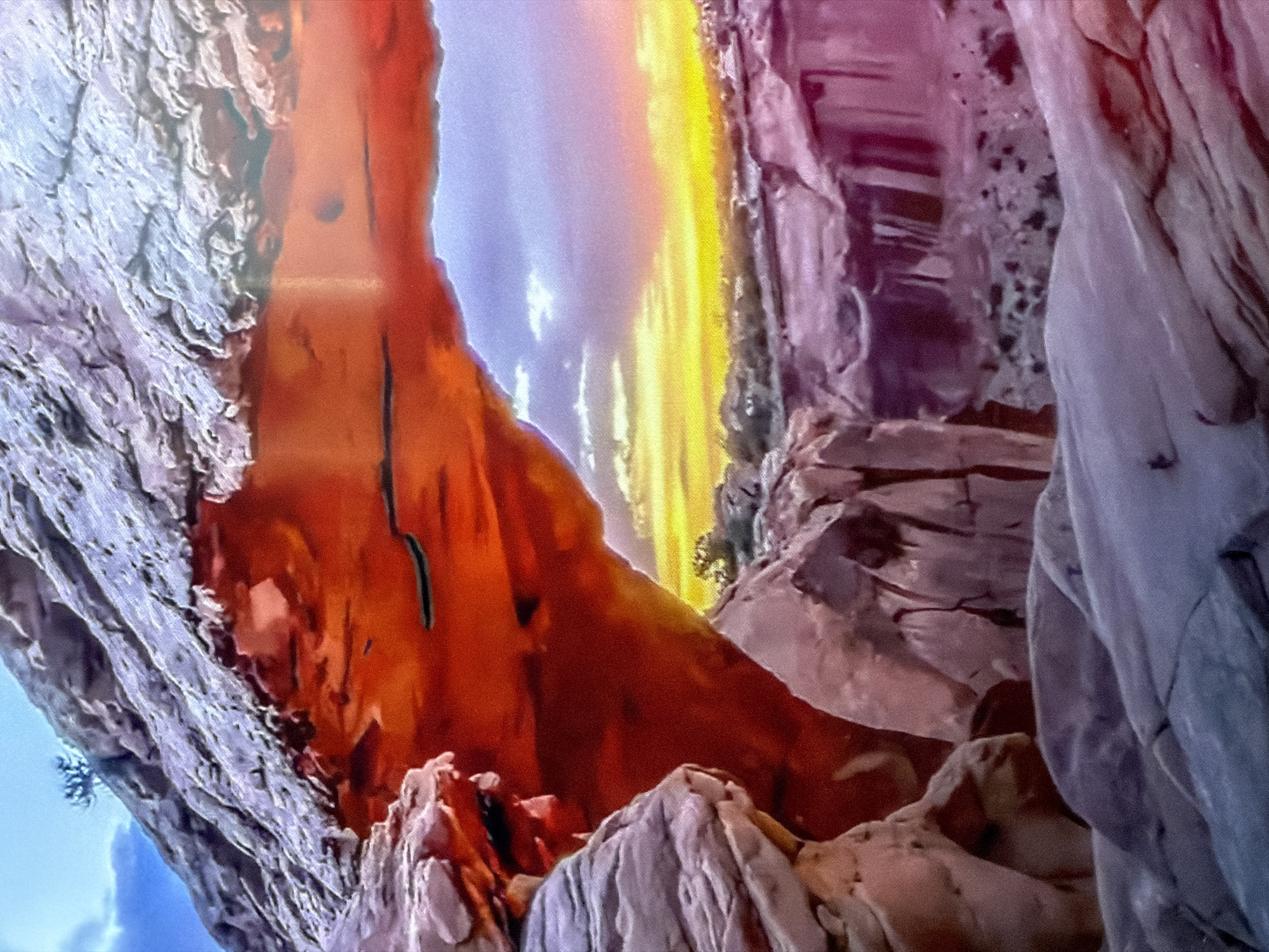 This is a landscape image featuring a dramatic canyon at sunset. The prominent subject is the natural geological formation, characterized by rugged rock faces and deep crevices. The canyon walls exhibit a range of colors, from pale grey and white on the left to warm oranges and reds in the center, transitioning to purples and darker tones on the right.
The activity depicted is a natural scene at sunset, with the sky displaying vibrant hues of yellow, orange, and purple, indicating the sun is low on the horizon. The light casts a warm glow on the rock formations, highlighting their textures and contours. There are no people or man-made objects visible in the image, emphasizing the raw beauty of the natural landscape.
Notable details include the harsh, weathered texture of the rocks, suggesting a dry and arid environment. The dramatic lighting and the deep shadows within the canyon create a sense of awe and solitude. The time of day is clearly sunset, and the weather appears clear to partly cloudy, with the sky showing a gradient of colors. The location cues point to a desert or mountainous region with significant erosion. There is no visible text in the image. The overall mood evoked is one of natural grandeur and peacefulness.
