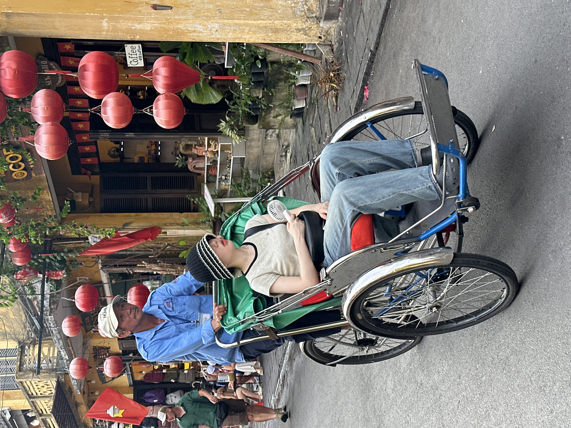 The image depicts a street scene in Hội An Ward, Vietnam, characterized by a weathered yellow building and a grey asphalt street. Red lanterns are strung across the street, adding a festive touch.
The central focus is a cyclo, a three-wheeled vehicle propelled by a rider. A man in a blue shirt and a white cap is operating the cyclo, facing forward with a neutral expression. Seated behind him, in the passenger compartment, is a young person wearing a light-colored t-shirt and a black and white striped beanie. This passenger appears to be resting or sleeping, with their head tilted back and eyes closed. A small white fan is clipped to the side of the cyclo, suggesting a warm day. The passenger is wearing blue jeans.
In the background, several shops are visible, adorned with flags featuring a yellow star on a red background, likely Vietnamese flags. One shop has a sign that reads "COFFEE." There are also shelves displaying various items, possibly souvenirs or goods for sale. The overall impression is a snapshot of daily life in a bustling tourist area, with the cyclo serving as a mode of transportation and a symbol of the local culture. The lighting suggests it is daytime.