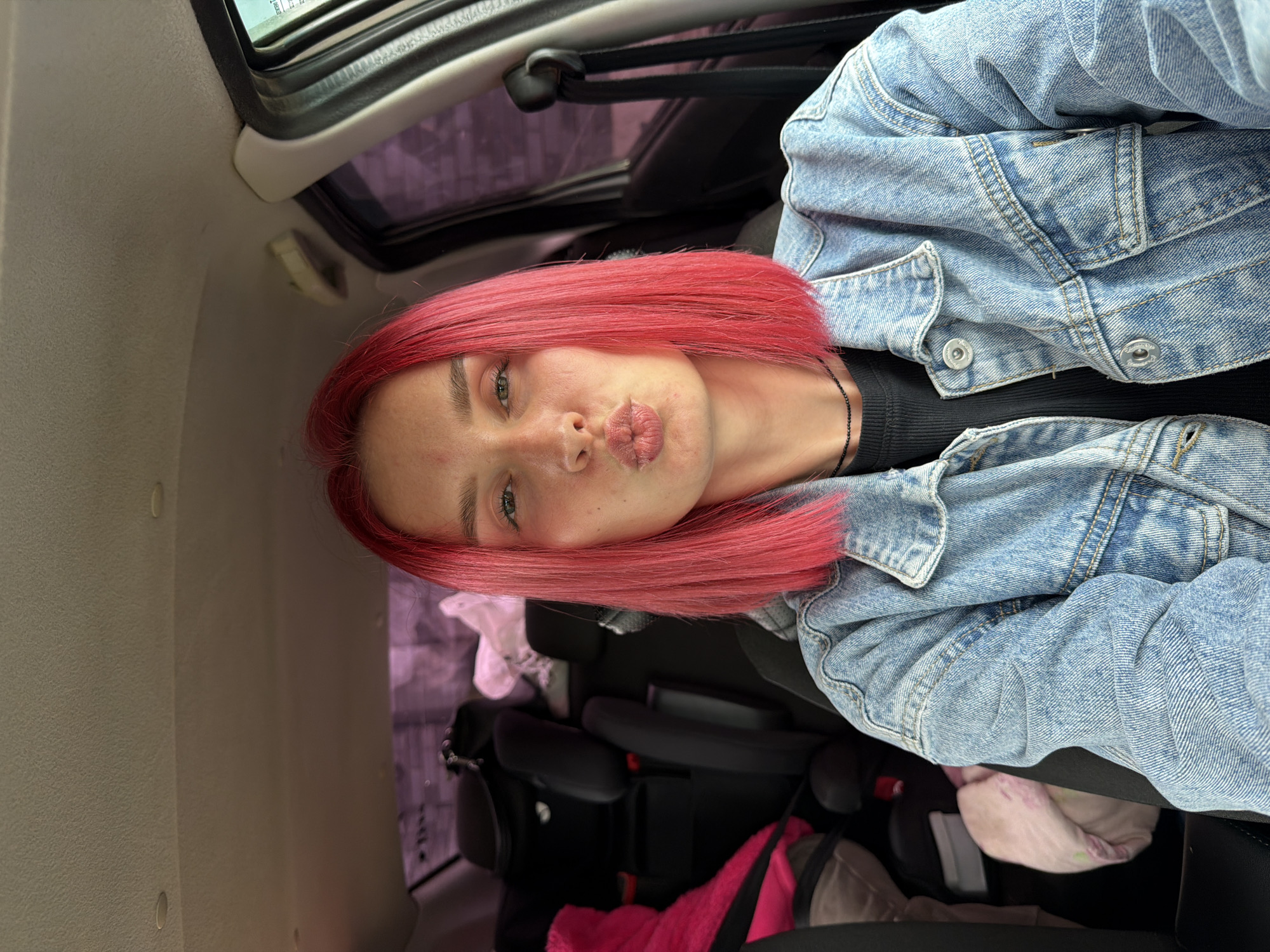 A young individual with short, bright pink hair, light eyes, and fair skin is depicted from the chest up, seated in the front passenger seat of a vehicle in Chaikovsky, Russia. The subject is wearing a light blue denim jacket over a black ribbed top and a black beaded necklace, and is making a puckering "kissing" expression towards the camera. The car's interior features a light beige headliner with two recessed dome lights. In the rear of the vehicle, two black child safety seats are visible, with a pink fabric item draped over the left seat and a light-colored fabric item on the right. The exterior environment, visible through the car's tinted windows, appears as a blurred urban or suburban landscape with structures in the background.