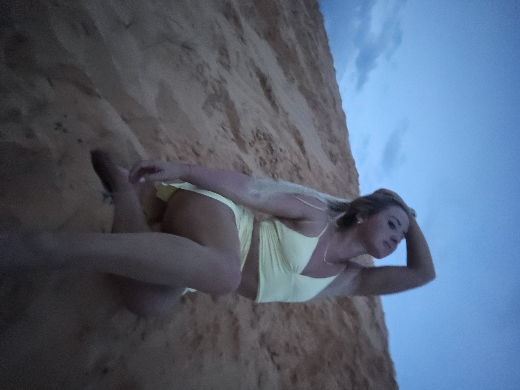 A young woman in a light yellow bikini poses on a sand dune in Mui Ne, Vietnam. She is lying on her side, with her head propped up by her hand. The sand is a light tan color and appears to be dry and loose. The sky is a clear blue with a few scattered clouds. The lighting suggests it is late afternoon or early evening. The woman has blonde hair and is looking off to the side with a thoughtful expression. The scene is peaceful and serene.