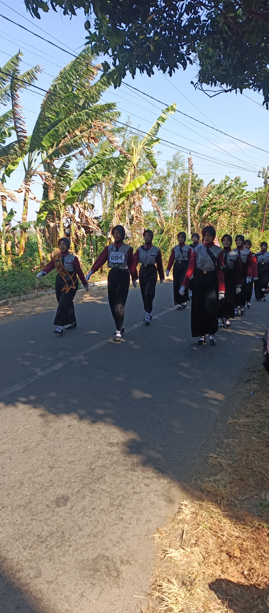A group of young people in uniform are marching down a paved road lined with trees and vegetation. The group appears to be a marching band or a similar organized group, as they are dressed in matching outfits consisting of maroon long-sleeved tops with gray collars and black pants. Several individuals are wearing black headscarves, suggesting a religious or cultural context. Some members have sashes or bibs with numbers on them, indicating they are part of a competition or event. The foremost individual on the left has a sash with text that is partially visible and appears to say "AL FALAH." The sky is clear and blue, and the sun is casting shadows on the road, indicating it is daytime. The environment suggests a rural or semi-rural setting, possibly in Indonesia, given the tropical vegetation like banana trees. The individuals are walking in formation, with their arms held out to their sides or in front of them, in a disciplined manner. The overall scene captures a moment of organized movement and community activity in an outdoor environment.
