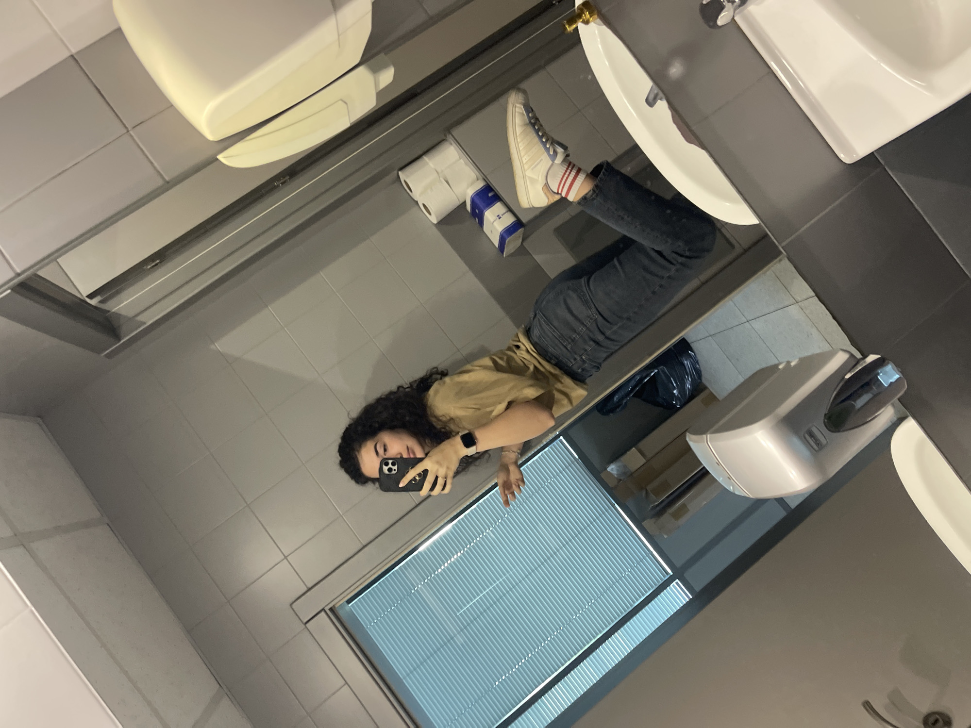 A mirrored selfie taken in a bathroom, likely a public restroom. The reflection shows a woman with curly hair holding a smartphone, taking a picture in front of a window with closed blinds. She is wearing a tan shirt and dark jeans. Her left leg is propped on the edge of the sink. The bathroom features white tile walls, a white sink, a toilet paper dispenser, a soap dispenser, and a hand dryer. The presence of Cyrillic letters on the toilet paper packaging suggests the location is in Bulgaria, with Sofia being the most likely city due to the larger population and higher probability of international brands.
