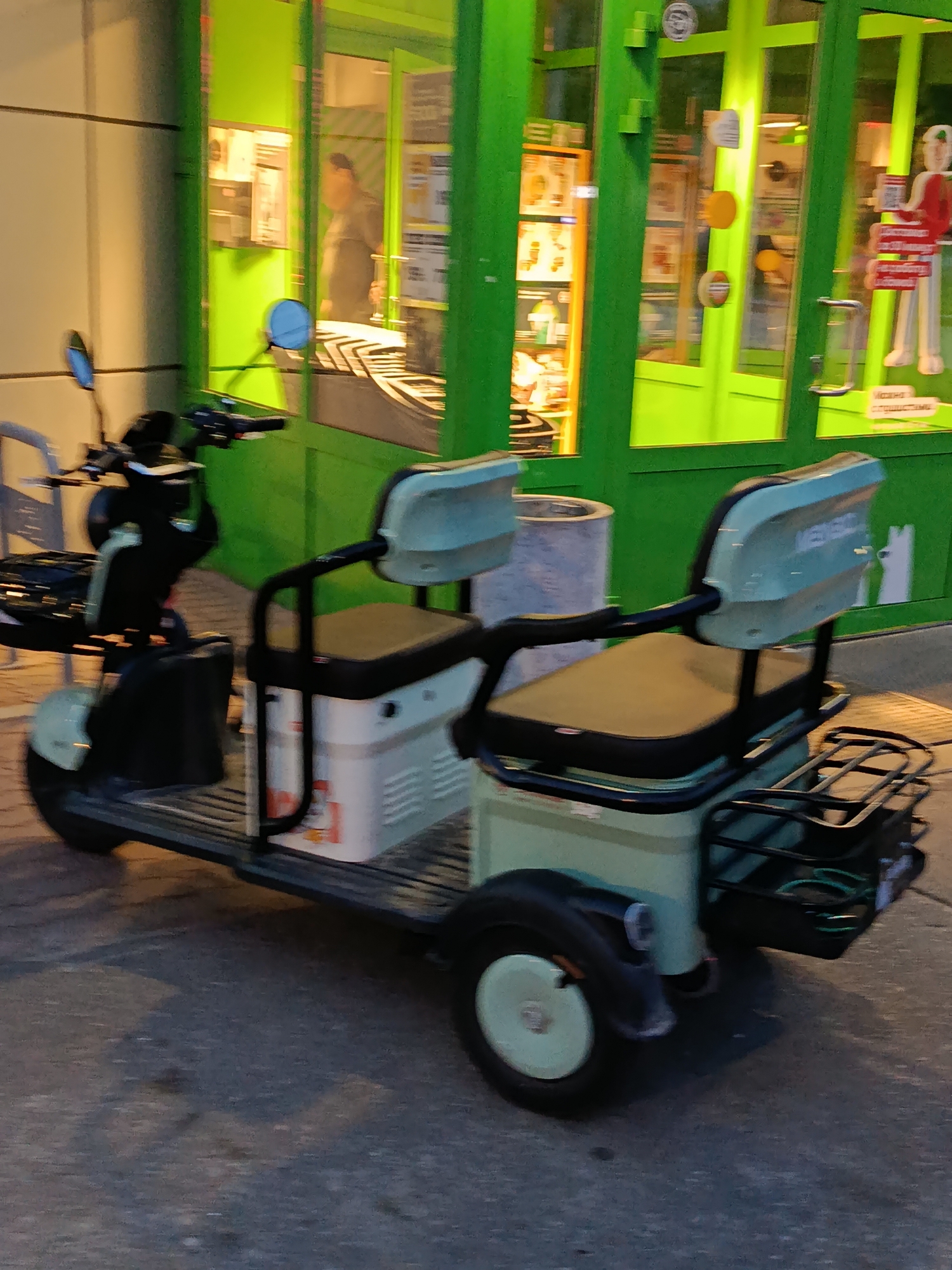 A three-wheeled electric scooter is parked on an asphalt surface in front of a bright green building with large glass windows. The scooter has two seats, a black frame, and light green body panels. It is equipped with a front basket and a rear rack. Inside the building, shelves are visible, stocked with various items, and a person is seen through the glass. The overall lighting suggests it is either dusk or the interior of the building is brightly illuminated. The location is identified as Verbya, Russia.