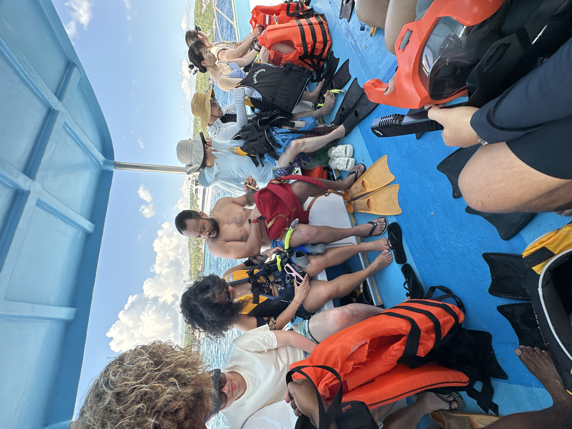The image shows a group of people sitting on a blue boat, presumably preparing for a water activity. Many are wearing life jackets or holding snorkeling gear such as fins and masks. Some are dressed in swimsuits or light clothing, suggesting a tropical environment. The background reveals a coastline with trees and buildings, suggesting a coastal region, consistent with Kampung Toyapakeh, Indonesia. The boat's interior is visible, with the blue hull and seating arrangement adding context to the activity taking place.
