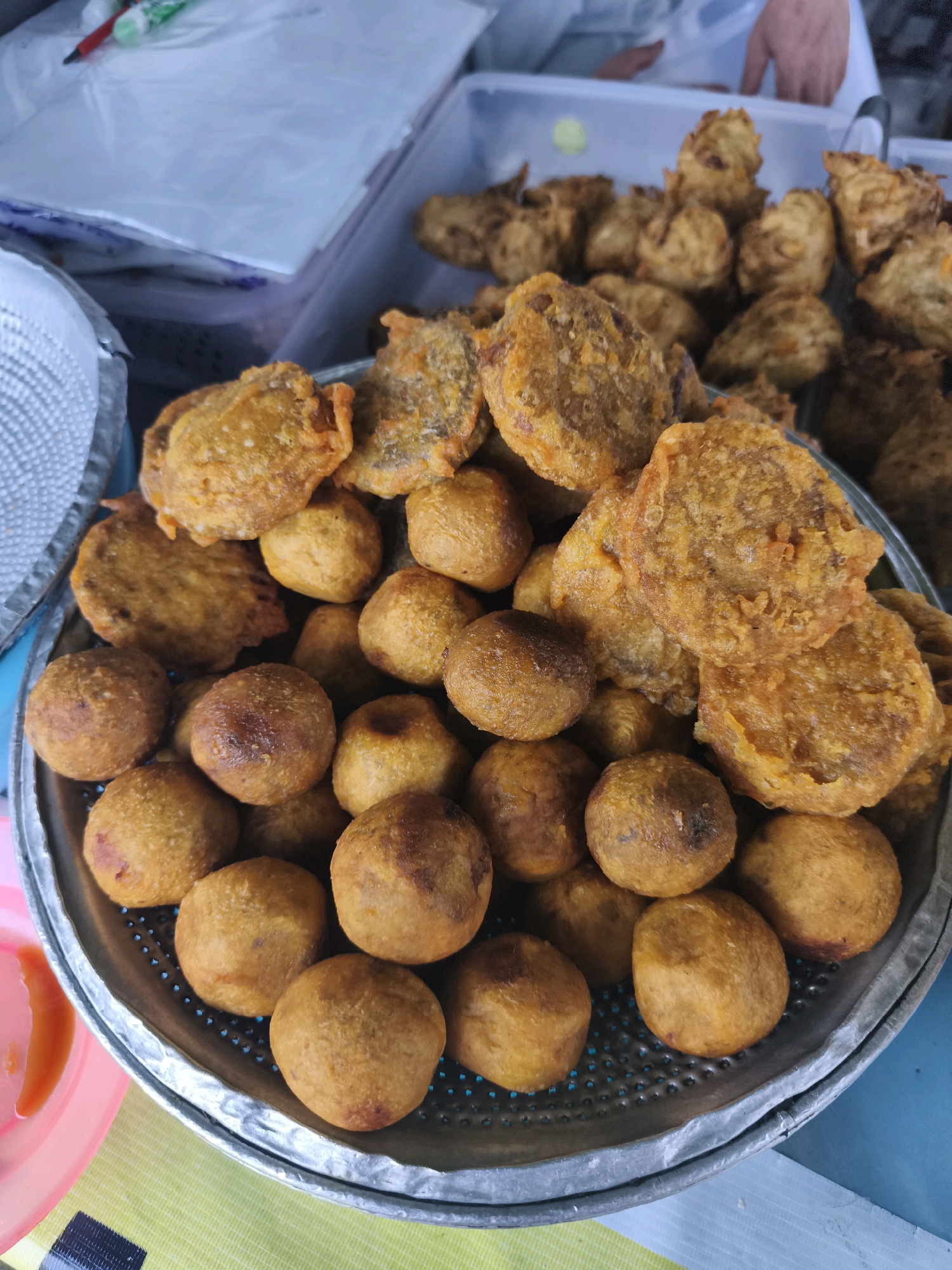 A close-up, overhead shot features a circular metal sieve filled with fried snacks. The snacks are of two types: round, golden-brown balls, and flatter, irregularly shaped fritters with a textured, slightly darker exterior. Both types appear to be made from a dough that has been deep-fried. The sieve is resting on a yellow-green checkered surface. In the background, a white plastic container holds more of the fried snacks, and another plastic container with a light blue lid is visible. A hand is partially visible in the upper right corner, reaching into the white container. To the left of the sieve, a pink plastic plate with a hint of orange sauce is visible. The overall setting suggests a food stall or market environment, possibly in Palembang, Indonesia, given the commonality of such fried snacks in Indonesian cuisine.