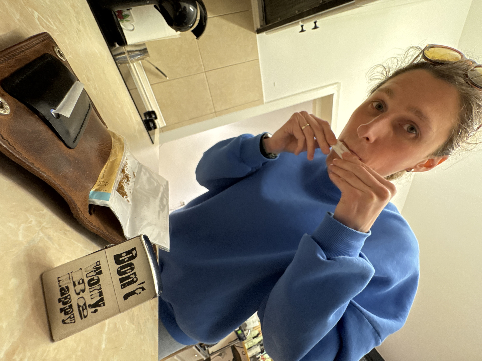 The image shows a woman with sunglasses on her head, wearing a blue sweater, and holding a cigarette in her mouth, in what appears to be an indoor setting. The background reveals a kitchen area with light-colored walls and tile flooring. On the counter, there is a brown leather case with a bag of tobacco and a silver lighter with the words "Don't Worry, Be Happy" printed on it. There is no information available in the image about Budva, Montenegro.